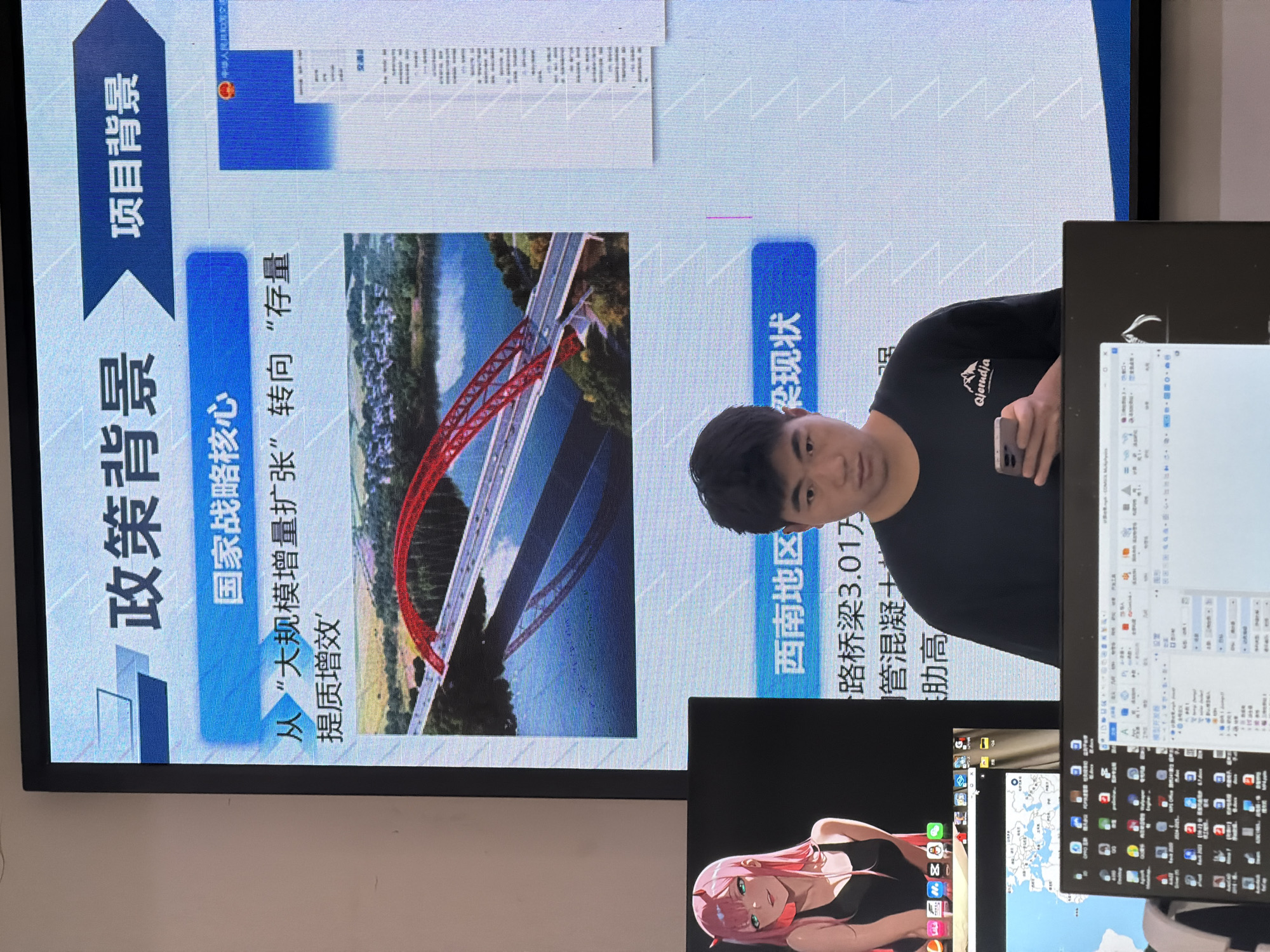 A person stands in front of a large screen displaying presentation slides. The screen shows Chinese text and images, including a red bridge over a body of water. The text on the screen translates to "Policy Background" and "Project Background." Below this, there is a section that reads "National Strategy Core" and "From 'Large-scale expansion' to 'Stockpile quality and efficiency'." Another section displays "Southwest Region" and "Infrastructure Status." The person is wearing a black t-shirt with a logo that says "Qierndja" and is looking at their phone. In the lower part of the image, multiple computer monitors are visible, displaying various windows and icons. One of the monitors shows a map. The lighting suggests an indoor environment, possibly an office or conference room. There is no visible indication of the city of Jiangjin, China.