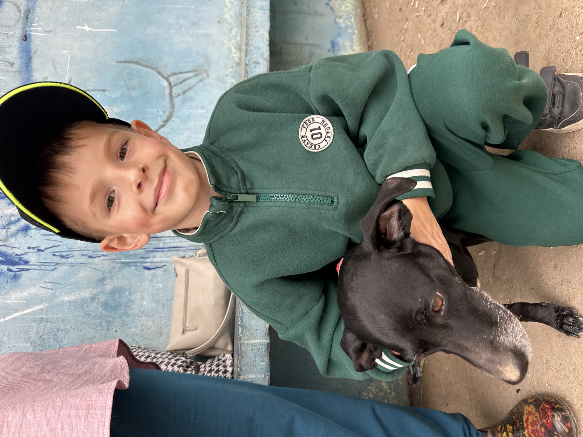 A young boy in a green tracksuit and black baseball cap sits next to a grey-muzzled black dog on a neutral-toned surface. The boy is smiling and has his arm around the dog. They are in what appears to be an urban area. Альняш, Russia is specified as the location. A light-colored handbag rests near the boy. A person wearing a pink shirt and teal pants is also partially visible.
