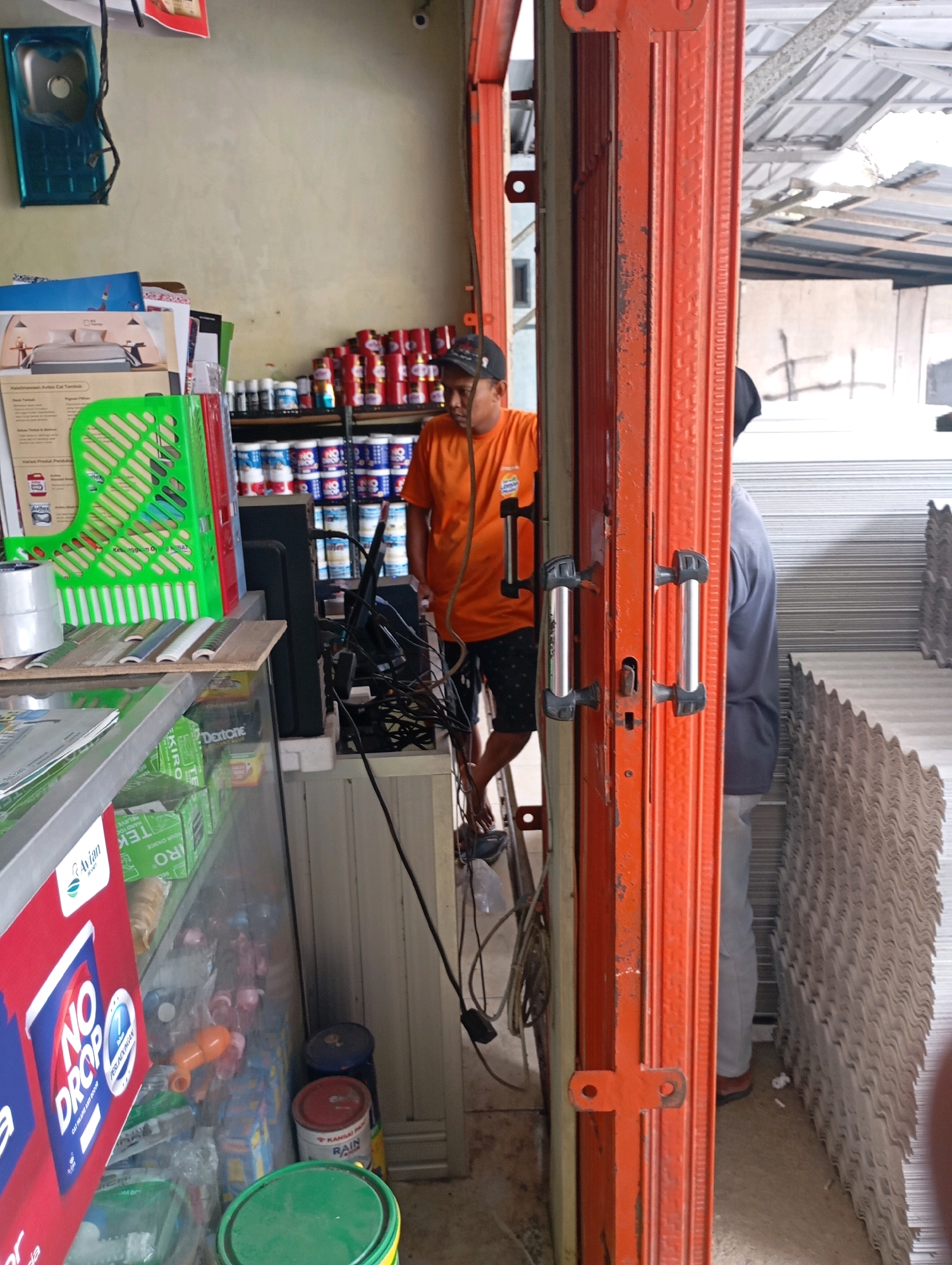 The image depicts a storefront in Wonogondo, Indonesia. A man in an orange t-shirt and patterned shorts stands inside the shop, leaning slightly. He is facing towards the right side of the frame, partially obscured by an orange metal gate. Behind him, shelves are stocked with various products, including cans of paint. Another person, wearing a grey t-shirt and light-colored pants, stands just outside the shop to the right, facing inwards. The environment appears to be an indoor or semi-outdoor space, with corrugated metal roofing visible in the background. The storefront features the orange metal gate with silver handles, which is slightly ajar. On the left side of the image, a blue sink is mounted on a wall, and a green plastic basket is positioned on a counter. The overall scene suggests a retail environment, possibly a hardware or paint store.