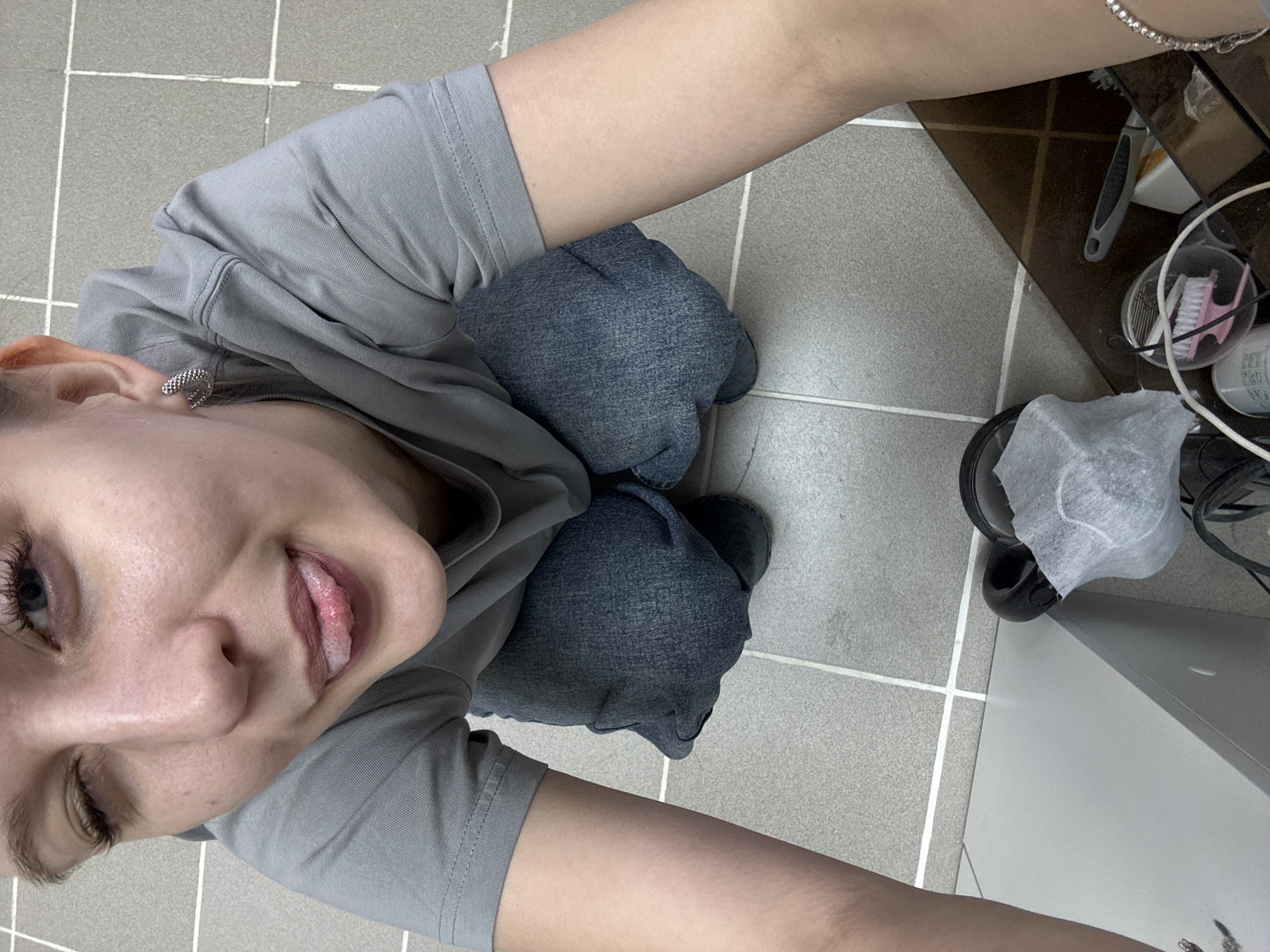 The image depicts a person with fair skin and blonde hair, wearing a grey t-shirt and blue jeans. The person is sitting on a tiled floor and looking at the camera. A trash can with a tissue sits on the floor next to a table. The table has personal grooming items on it. The location is unknown, but inferred to be in Izhevsk, Russia. No additional details are available.