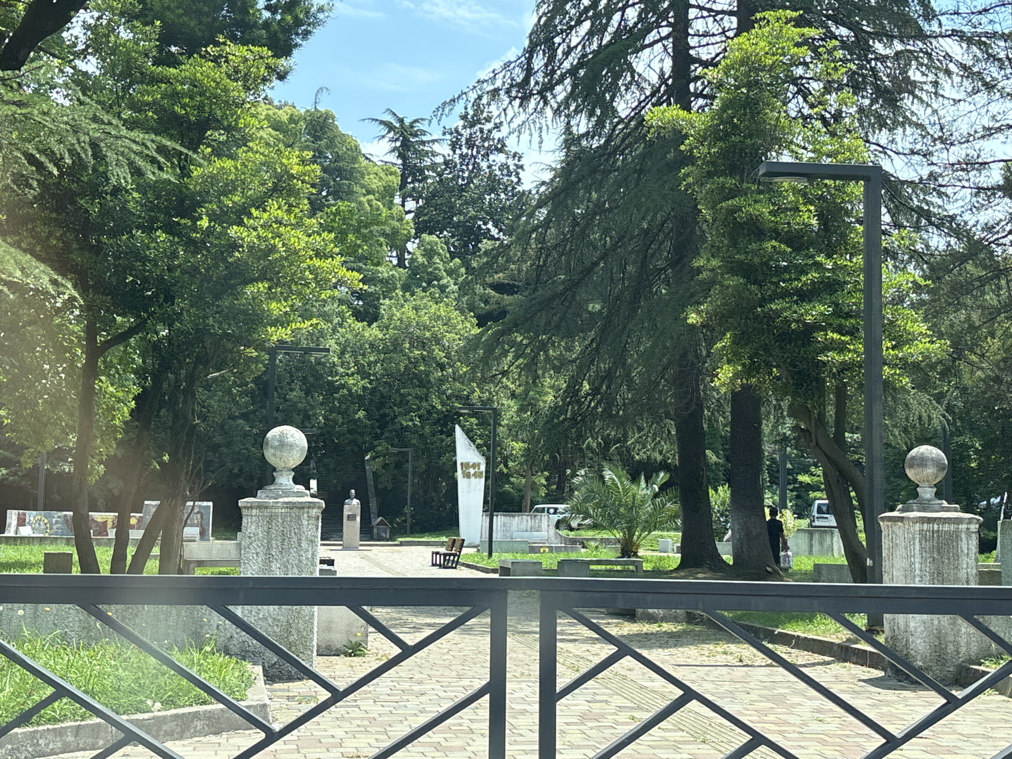 The image depicts a public park area in Batumi, Georgia, on a sunny day. In the foreground, a black metal fence with a geometric pattern separates the viewer from the park. Beyond the fence, a paved walkway is visible, with an ornate stone pillar topped by a spherical finial to the left and another similar pillar to the right. A lush green lawn with grass and some scattered yellow wildflowers is also present.
In the middle ground, a white obelisk-like monument with gold lettering "1941 1945" stands prominently. Adjacent to it are two wooden benches. Further back, there are trees, some large and mature with dense foliage, and a smaller palm tree. A bust on a pedestal is visible to the left of the monument, with a set of stairs leading up behind it. A white vehicle is parked in the background. A person wearing dark clothing is walking near a tree on the right side of the frame, carrying what appears to be a white bag. The background is filled with a variety of trees, creating a green canopy, with a bright blue sky with scattered white clouds overhead. The overall impression is of a well-maintained park with historical markers.