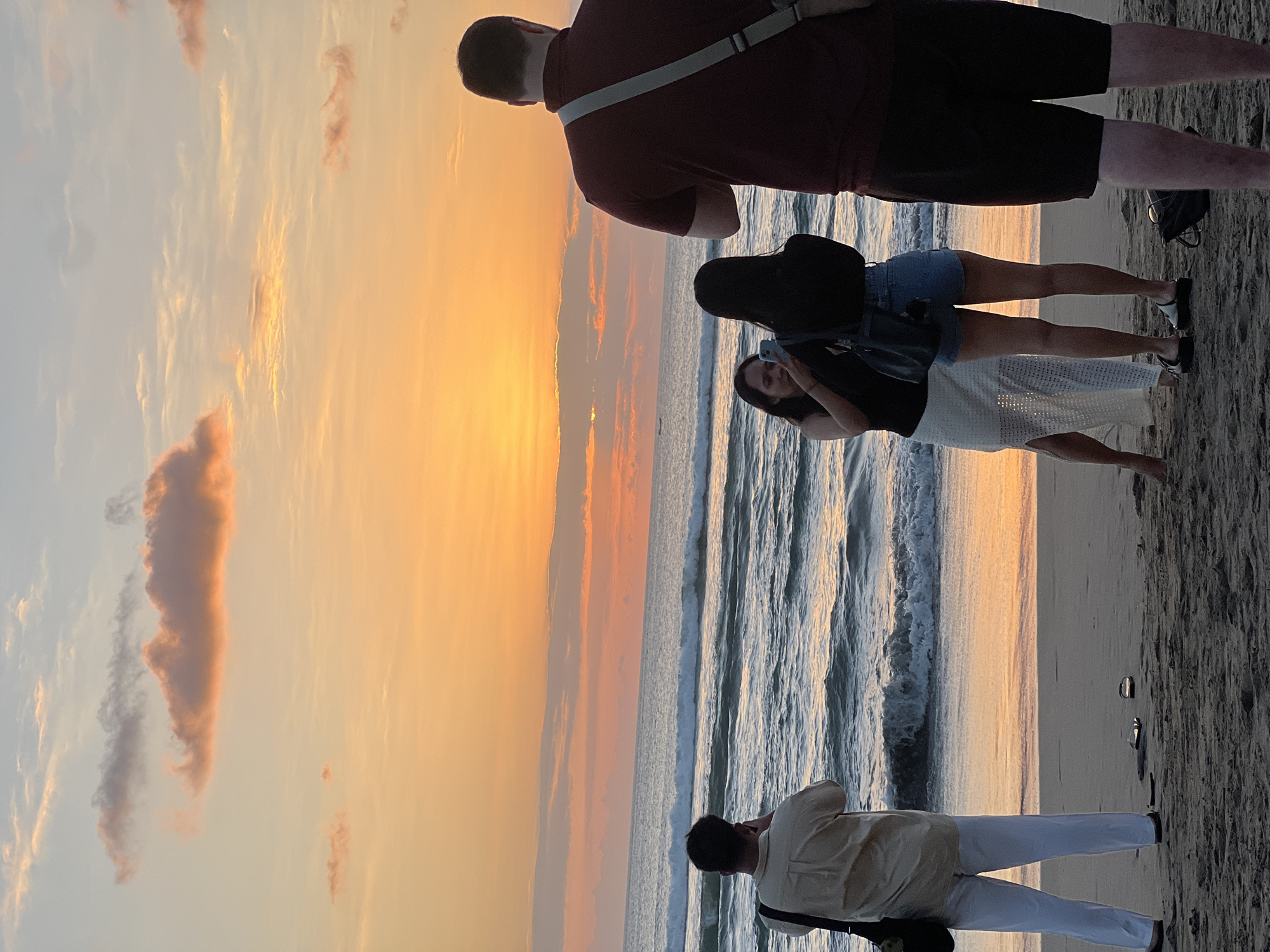 This image depicts a sunset scene on Kuta Beach, Indonesia. Four individuals are present on the sandy shore, facing the ocean. In the foreground, a person on the far right, facing away from the camera, wears a dark red polo shirt and dark shorts. To the left, a person in a light-colored short-sleeved shirt and white pants stands with a dark bag slung over their shoulder, holding a phone to their ear. In the middle ground, two women are engaged with a smartphone; one, wearing a black top and a long, white, semi-sheer skirt, holds the phone while the other, in a black top and denim shorts, is near her. The ocean in the background features moderate waves, and the sky is painted with vibrant orange and pink hues from the setting sun, with scattered clouds. The beach is composed of dark sand.