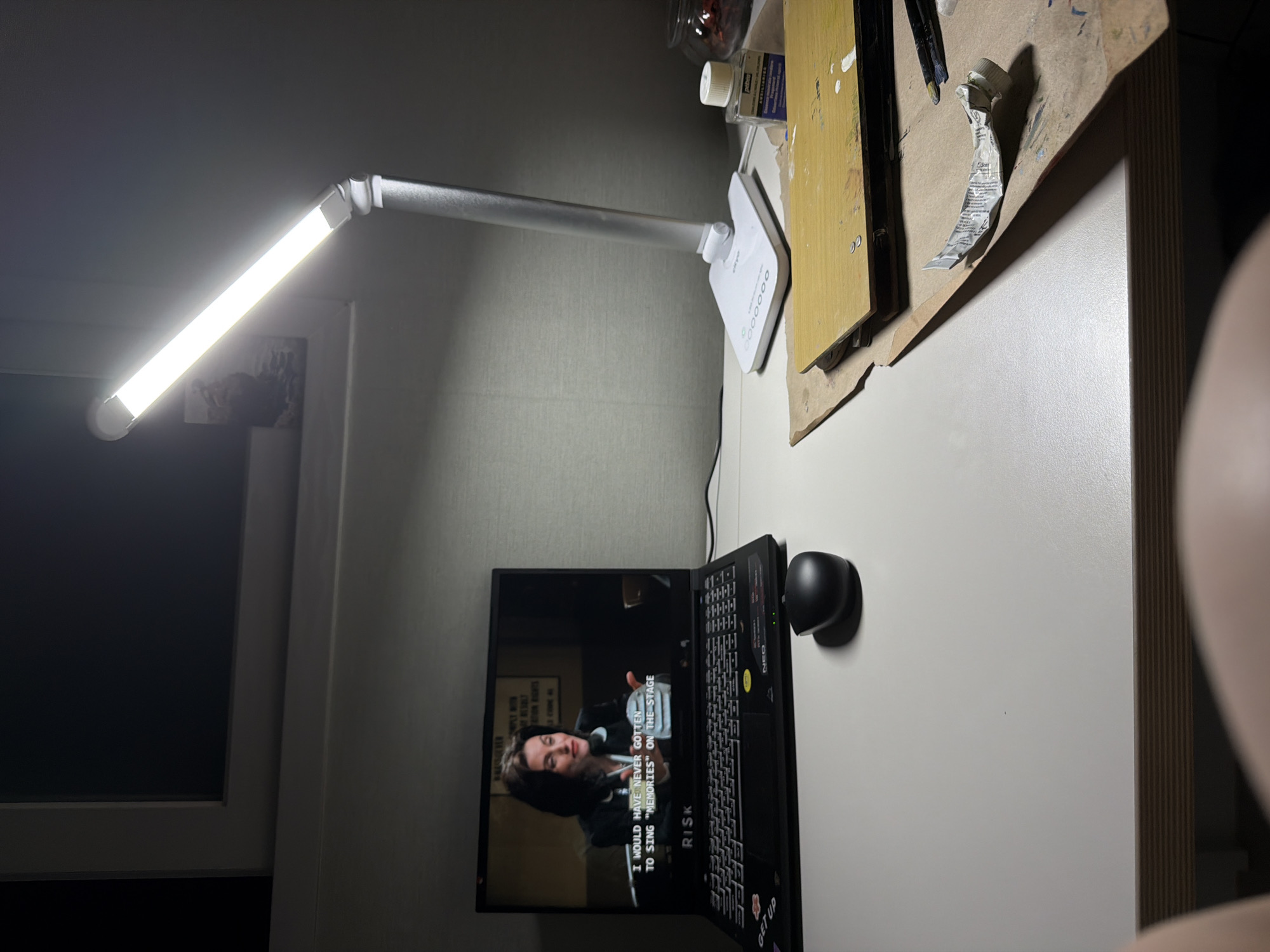 The image shows a desk setup with a laptop open and a lamp providing light. The laptop screen displays a person speaking with subtitles, possibly from a video or movie. The subtitles read: "I WOULD HAVE NEVER GOTTEN TO SING 'MEMORIES' ON THE STAGE". The laptop is black and has various stickers on its surface, including one with the word "RISK" and another with "GET UP". A small black computer mouse is positioned to the right of the laptop.
A silver LED lamp with a long, thin light bar is positioned above and to the left of the laptop, illuminating the desk. Behind the lamp, on the desk, are art supplies, including a wooden palette with paint smudges, tubes of paint, brushes, and a jar. The background appears to be a wall with a textured surface. The overall scene suggests a creative workspace or a personal study area, likely indoors. The lighting suggests it might be evening or a dimly lit room. The location is specified as Dongducheon-si, South Korea.