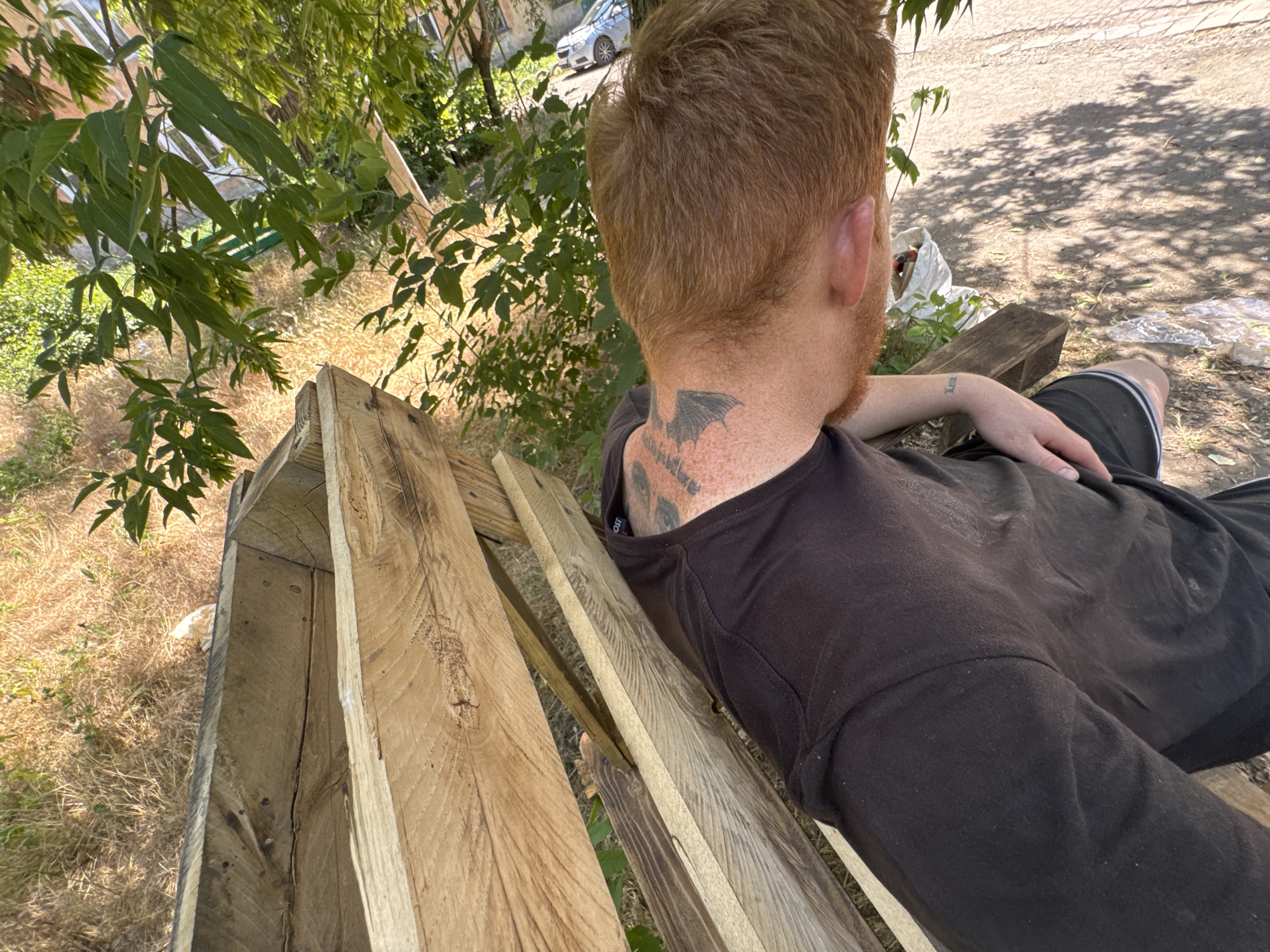 The image shows a person sitting on a wooden bench outdoors. The person has reddish hair and a tattoo on the back of their neck which appears to be a bat. They are wearing a dark t-shirt and dark shorts with white stripes. Their left arm is resting on their leg, and a small tattoo is visible on their wrist.
The bench is made of rough, weathered wooden planks. Behind the bench, there is dry, yellowed grass and some green foliage from trees overhead. In the background, a paved area is visible with tire tracks and a car parked in the distance. Sunlight filters through the trees, casting shadows on the ground. There is a white plastic bag and some other litter scattered near the bench. The overall scene suggests a casual, possibly neglected outdoor space on a sunny day. The lighting indicates it's daytime, likely mid-morning or afternoon due to the strong shadows. No discernible emotions are conveyed by the visible part of the person. There is no visible text in the image.