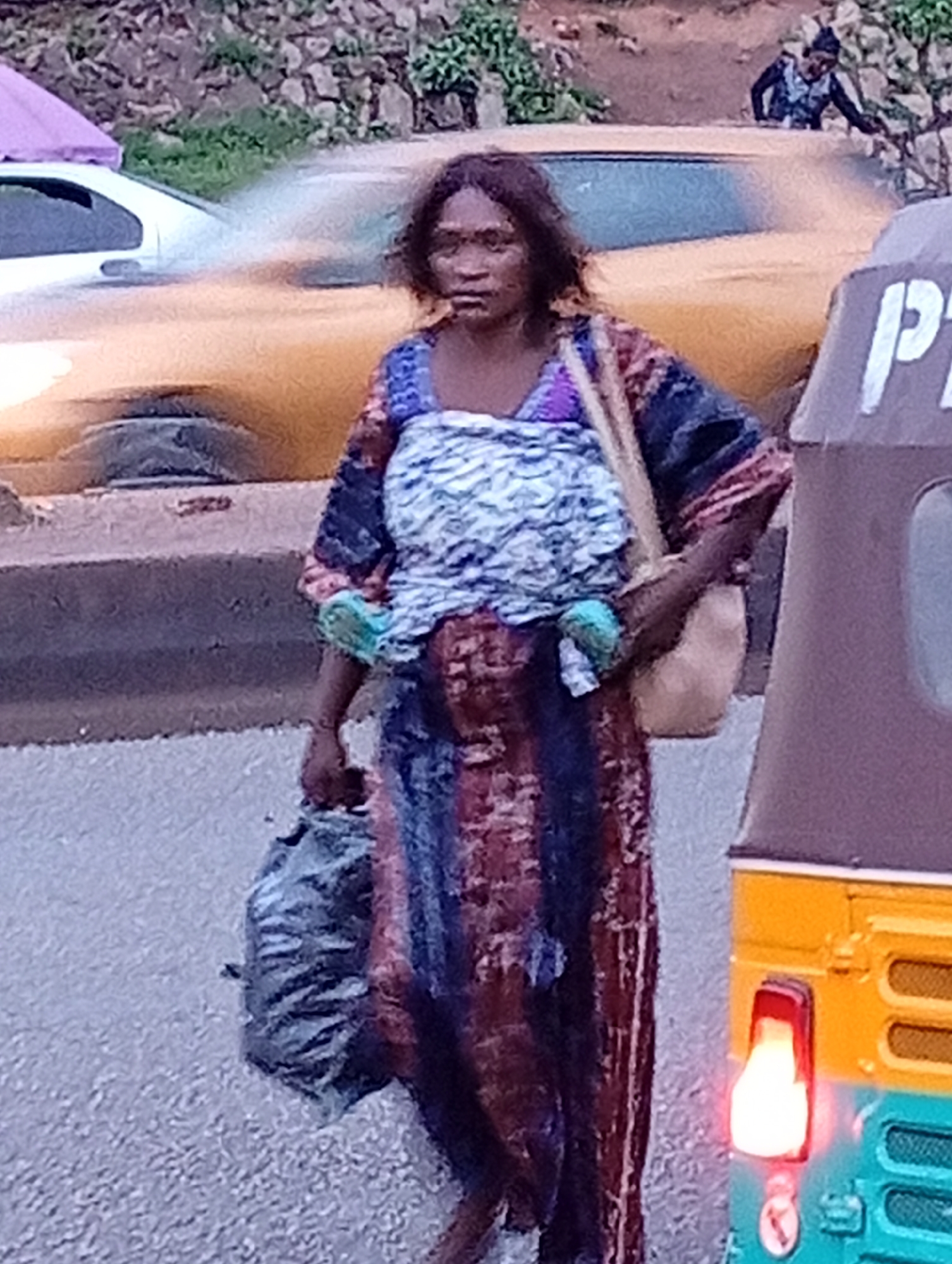 A woman stands by a street in Jos, Nigeria, near a yellow car and a multi-colored motorized rickshaw. She wears a patterned dress and carries a large bag in her left hand. The rickshaw has a red brake light illuminated. In the background, there are trees and a stone wall.
