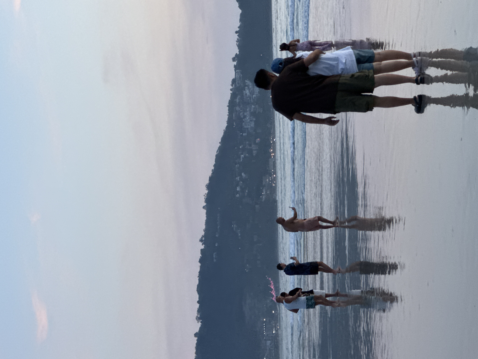A group of individuals are standing on a wet, reflective beach in Patong, Thailand, during twilight. The sky is a gradient of pale blue and pink. In the foreground, a couple stands facing away from the camera, with the man embracing the woman from behind. The man is wearing a dark t-shirt and olive-green shorts, and the woman is wearing a light blue cap and a white shirt with blue shorts. To their left, further down the beach, four other individuals are visible. One man in a white tank top and patterned shorts is holding a phone. Another man in a dark blue patterned shirt is also holding a phone. A third man, bare-chested, is holding a phone and appears to be taking a photo or video. Behind him, a woman in a light-colored outfit stands. In the distance, a tree-covered hill rises, with lights from buildings visible on its slopes. The sea is calm with small waves lapping the shore. The wet sand creates a mirror-like reflection of the people and the sky.