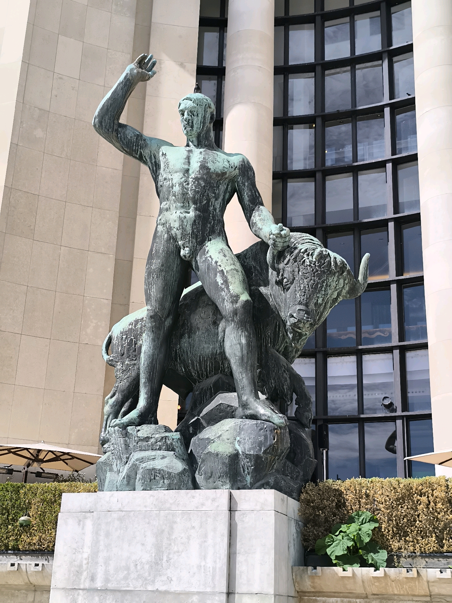 This is a frontal, medium-shot photograph of a large bronze statue of a muscular male figure riding a bull. The statue is situated outdoors in front of a modern building with a facade of light-colored stone and a large, multi-paned glass window. The bronze of the statue has a greenish patina, indicative of age and exposure to the elements.
The male figure is depicted with his left arm raised, hand open, and his right arm resting on the bull's back. His posture is dynamic, suggesting movement or exertion. The bull is powerfully built, with its head turned slightly towards the viewer, its horns prominent. Both the man and the bull are positioned on a rocky base, which itself rests on a large, light-colored stone pedestal.
In the background, to the left and right of the statue, are outdoor seating areas with white umbrellas and planters filled with greenery. The building behind the statue features a curved section of windows, reflecting the sky and surrounding environment. The lighting suggests it is daytime, possibly overcast given the muted colors and reflections in the windows.
This statue is likely one of the "La Défense de Paris" sculptures by Louis-Albert Carvin and Henri-Alfred Jaussely, located in the Trocadéro Gardens in Paris, France.