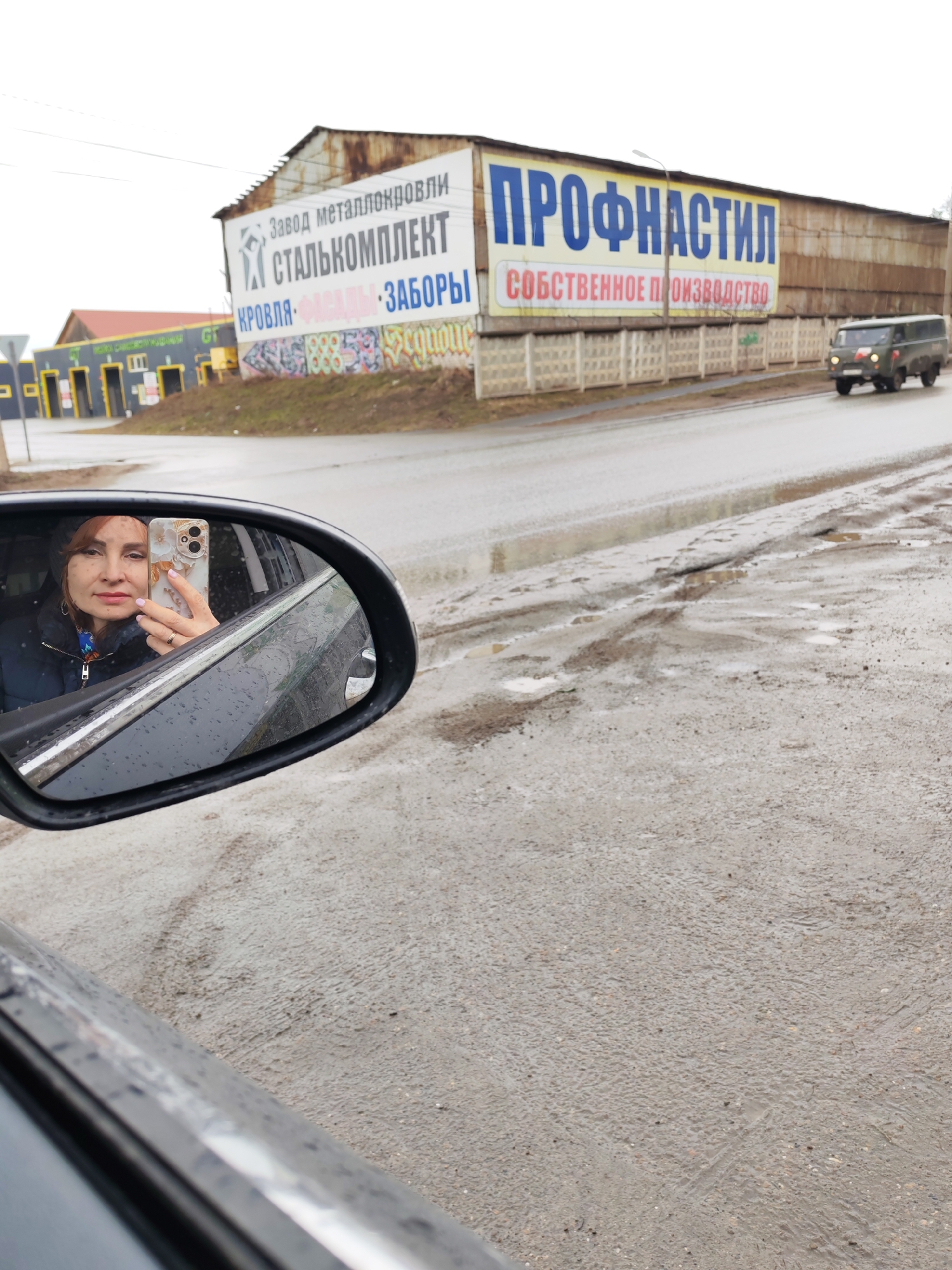 The image, taken from the driver's seat of a vehicle (likely a car) via the side-view mirror, presents a scene in what appears to be an industrial or commercial area. In the side-view mirror, a woman is visible, holding a smartphone and taking a picture of herself. She appears to be wearing a dark jacket and a hat.
The external environment shows a wet and potentially muddy road with puddles. On the other side of the road is an industrial building with large advertisements. One advertisement features the words "СТАЛЬКОМПЛЕКТ" (STALKOMPLEKT) and "КРОВЛЯ ФАСАДЫ ЗАБОРЫ" (ROOFS FACADES FENCES). Another sign on a larger building reads "ПРОФНАСТИЛ СОБСТВЕННОЕ ПРОИЗВОДСТВО" (PROFILE OWN PRODUCTION). An off-road vehicle appears to be moving on the road away from the camera.
Based on the Cyrillic signage, the location is likely within Russia, specifically Chaikovsky. The businesses advertised, dealing with metal products for construction (roofing, facades, fences), provide further clues about the area being an industrial zone.