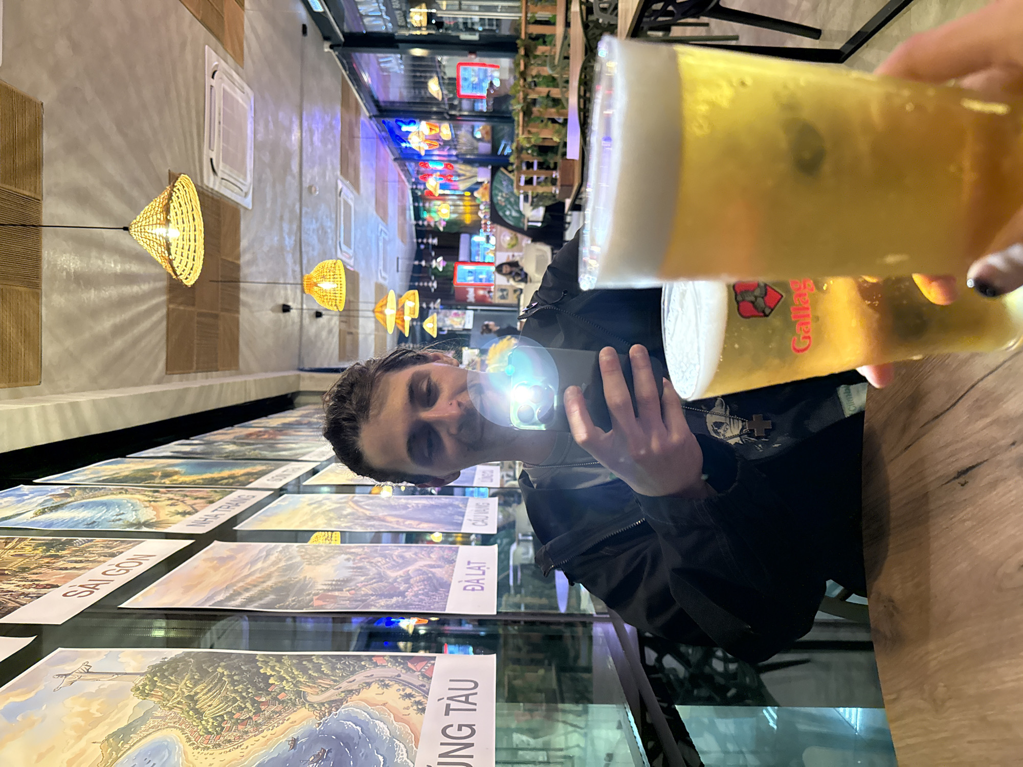 A man is holding a phone up to take a picture, in front of him is a table with picture postcards of different Vietnamese cities. The postcards, from left to right, show the cities of Vung Tau, Da Lat, Cau Vang, Nha Trang, and Saigon. Two glasses of beer are visible in the foreground. The background includes restaurant lighting and more seating. The location in the city of Sochi, Russia, is unknown from the image.