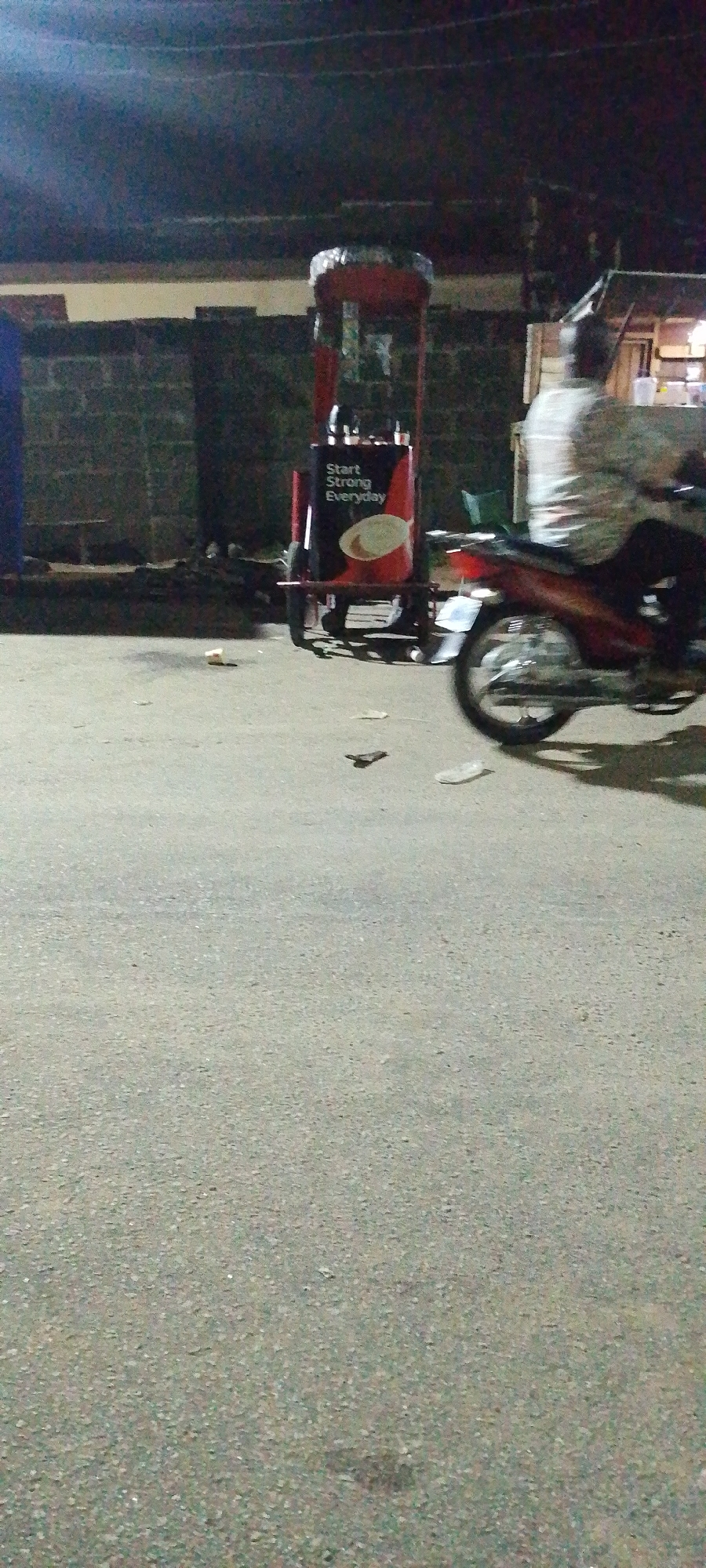 The image depicts a street scene in Lokoja, Nigeria, at night. The scene is illuminated by street lights. In the middle ground, there is a food vending cart with the slogan "Start Strong Everyday" written on its side. A person on a motorcycle is partially visible on the right, with only the rear portion of the bike and the person's back visible. The foreground is dominated by a paved road, with some scattered debris. A brick wall is visible in the background on the left. A small roadside shed is also visible on the right, behind the motorcycle.