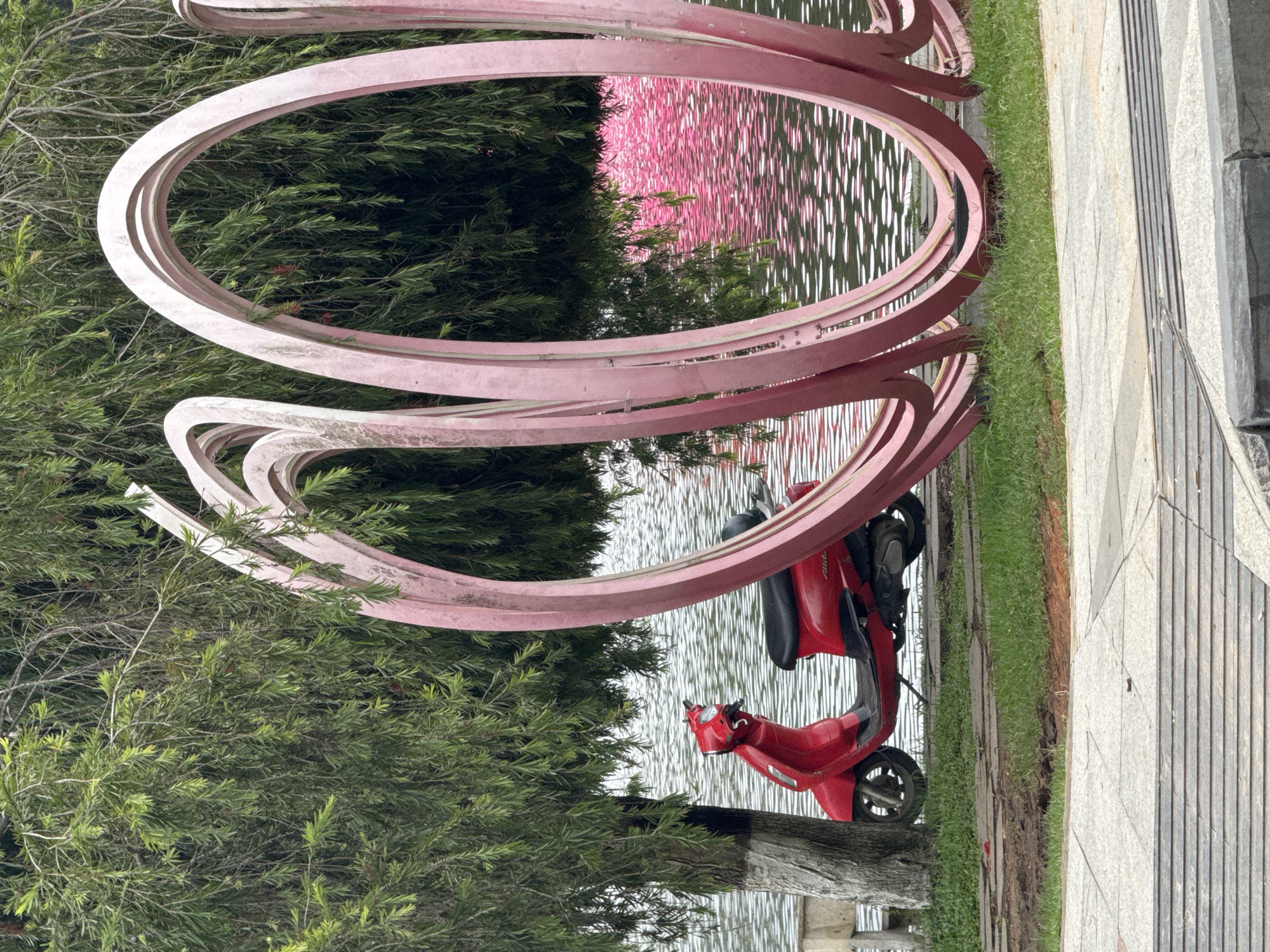 A red scooter is parked near a pink metal sculpture consisting of interconnected, concentric circles. The location is Da Lat, Vietnam, as the sculpture is a recognizable landmark of the city. The scooter is situated on a small patch of grass bordering a body of water. Reflections on the water are visible through the circles of the sculpture. A paved walkway is adjacent to the grass. Trees and greenery are present throughout the scene.