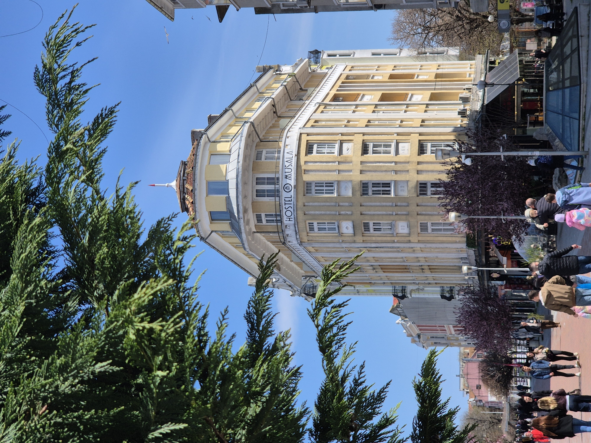 The image shows a building in Varna, Bulgaria, with the sign "Hostel Musala" on its facade. The building is multi-storied and painted in yellow and white. In the foreground, there are evergreen trees with dense foliage. The sky is clear and blue. The scene is a street view with pedestrians walking on the pavement. Some people are standing near the building.