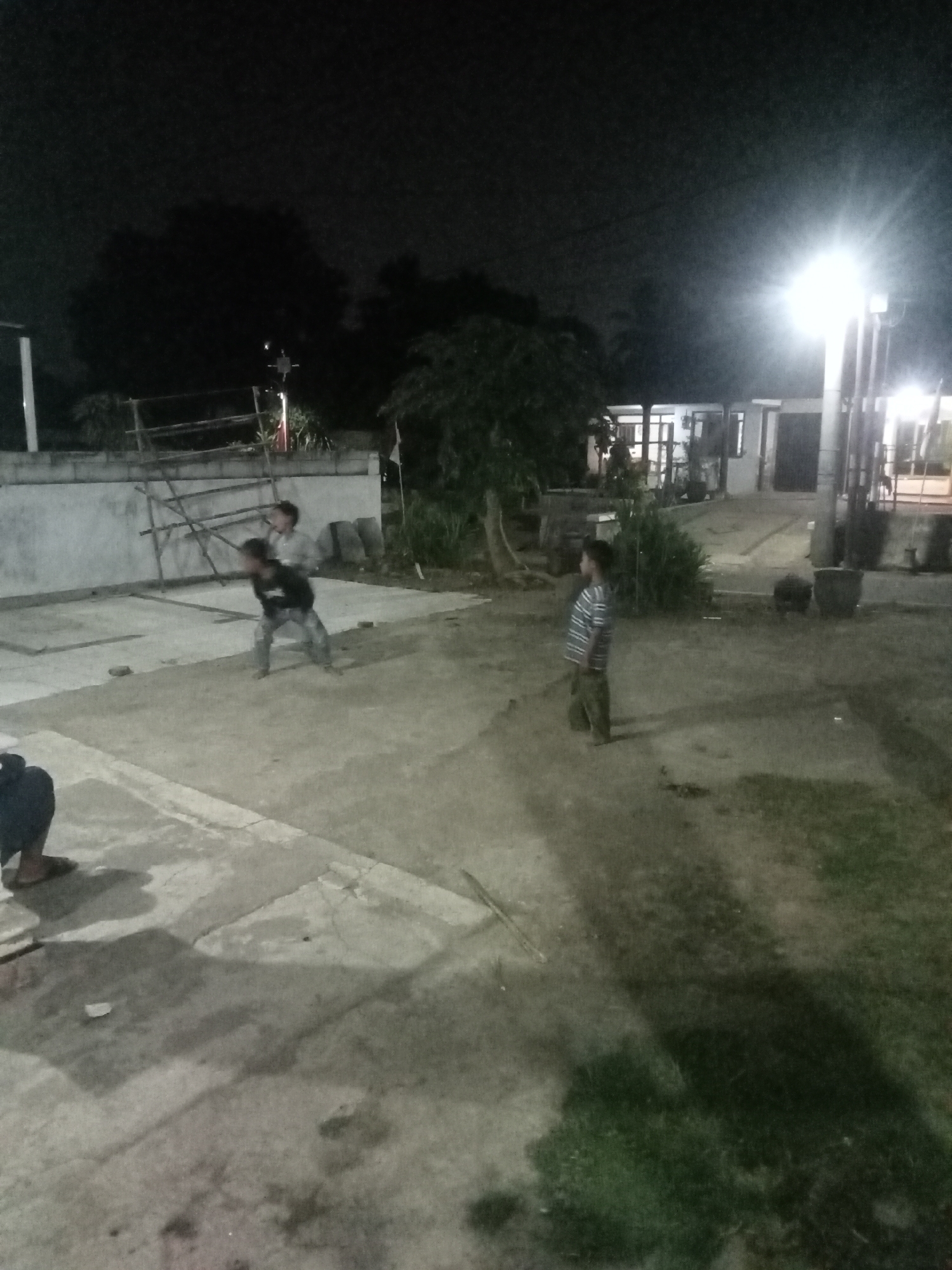 The image depicts a nighttime scene in an outdoor area, possibly a courtyard or open space in front of buildings. The lighting is dim, primarily from a bright overhead lamp on the right side of the frame and interior lights from the buildings in the background. There are at least three individuals visible. In the mid-ground, two young males are engaged in an activity, one in a dark shirt and jeans, appearing to be in motion, and another in a lighter-colored shirt behind him, also blurred by movement. To the right, a third young male in a striped shirt and pants stands facing away from the camera, looking towards the buildings. In the foreground on the left, a person's legs and feet, wearing sandals, are visible, suggesting they are seated or standing out of frame. The environment includes concrete pavement with some grass areas and shadows cast by the lighting. In the background, there are low-rise buildings with visible windows and structures that might be part of an ongoing construction or renovation project, like scaffolding. The overall impression is of a casual gathering or activity taking place in a somewhat urban or suburban residential setting at night. The location is identified as a city in Malang, Indonesia.
