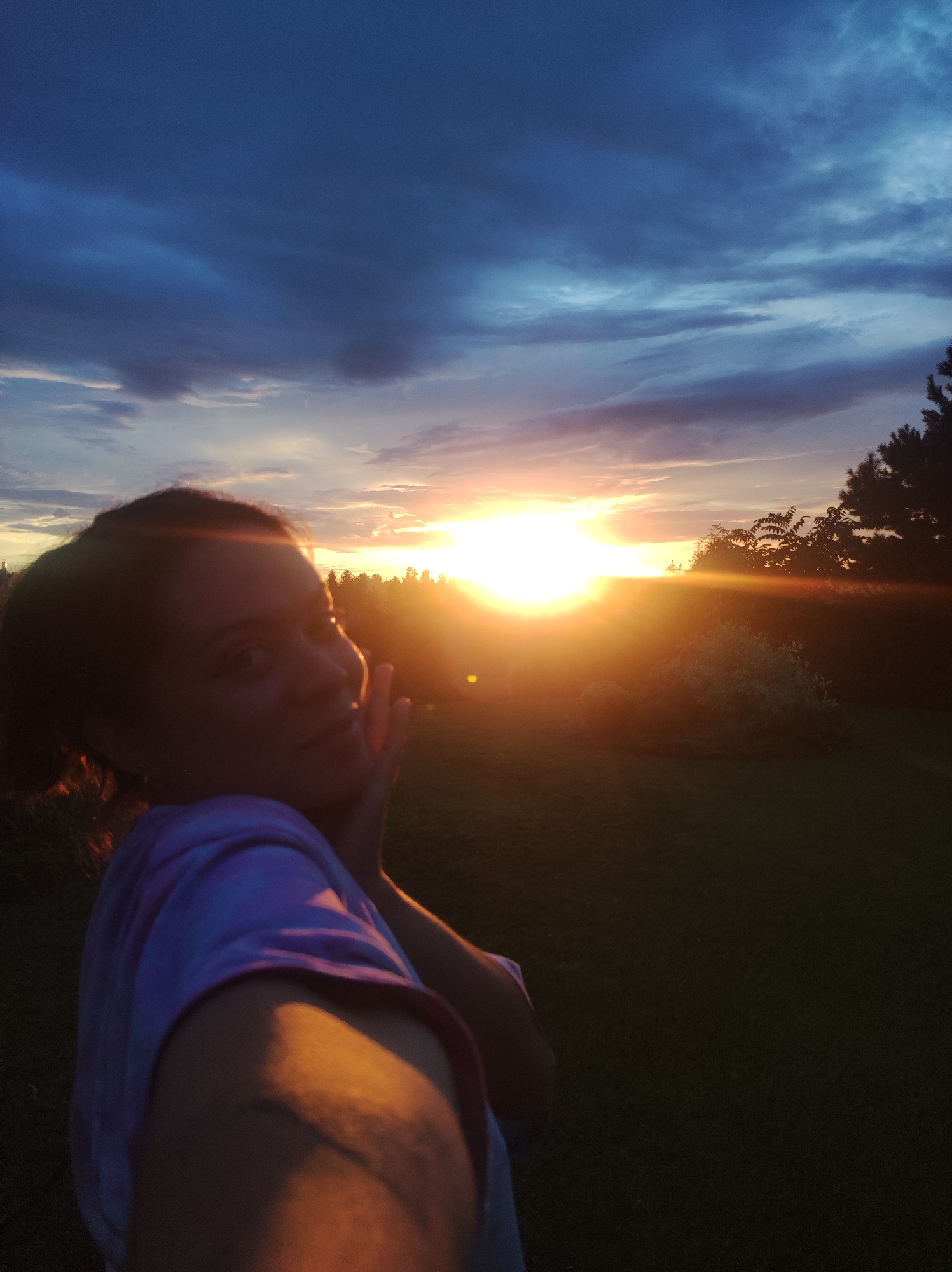 The image is a selfie taken outdoors during sunset, likely in a rural or suburban setting in the city of Fokino, Russia. The foreground features the upper body and face of a woman, seen from a slightly low angle. She has dark hair pulled back and is wearing a light-colored, possibly tie-dyed, hoodie or sweatshirt with purple and white hues. Her right hand is raised to her cheek in a posed gesture. The woman's face is illuminated by the setting sun, creating a warm glow.
The background is dominated by a dramatic sunset sky with a mix of deep blues, grays, and vibrant oranges and yellows. The sun itself is a bright orb, casting long rays of light and creating lens flare. Silhouetted against the sunset are trees and foliage, forming a dark line across the middle ground. Beyond the trees, the landscape appears to be a grassy field, mostly in shadow. The overall impression is one of serene natural beauty at dusk.