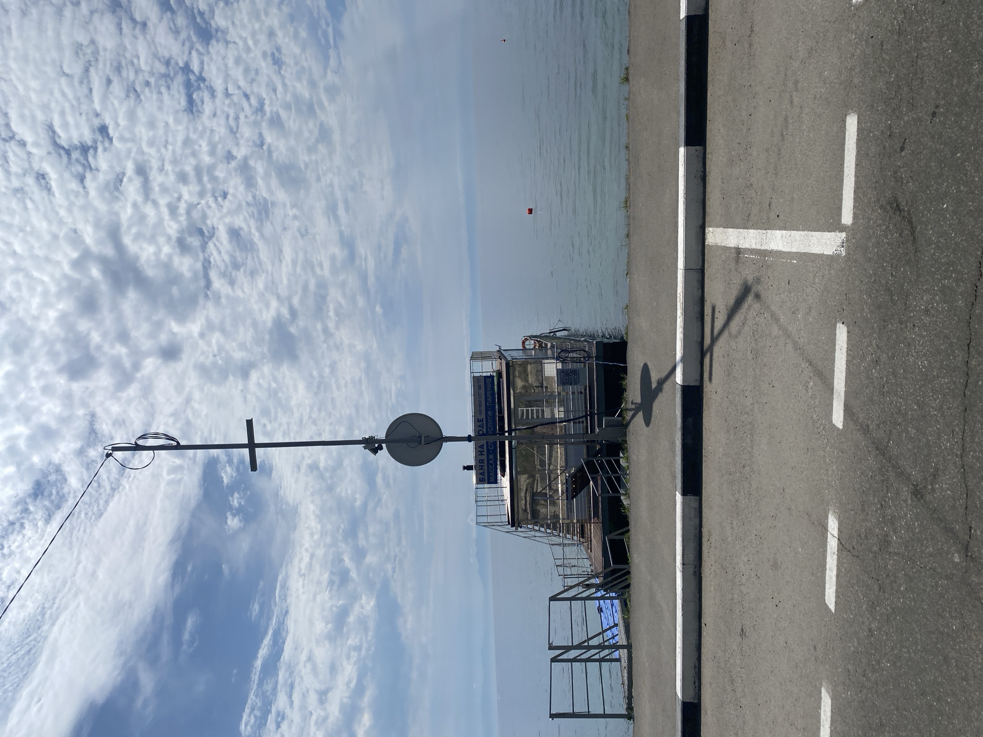 The image depicts a waterfront scene in Listvyanka, Russia, featuring a floating sauna on a calm body of water. In the foreground, a paved road with white parking lines is visible, bordered by a black and white curb. A tall metal pole with a circular sign and a crossbar stands near the curb. The floating structure, identified as a sauna by Russian text on its side, is accessible via metal stairs and a walkway. The water is calm, with a few buoys visible in the distance. The sky is partly cloudy with a mix of white and grey clouds against a blue backdrop. The overall scene is quiet and still, suggesting a peaceful day by the water.