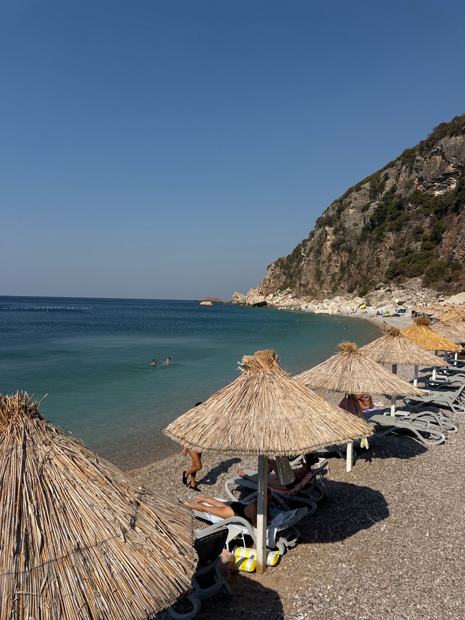 A sunny day at a pebble beach in Mirište, Montenegro, features several thatched umbrellas providing shade over empty lounge chairs. In the foreground, the edge of a large thatched umbrella is visible. The sea is a clear, turquoise blue, with two people swimming in the distance. A rocky, tree-covered cliff face rises steeply from the beach on the right side of the image, and a faint line of buoys stretches across the water towards the horizon. Several people are visible on the beach and in the water, and there are parked vehicles further down the shore. The overall impression is one of a relaxed coastal vacation setting.