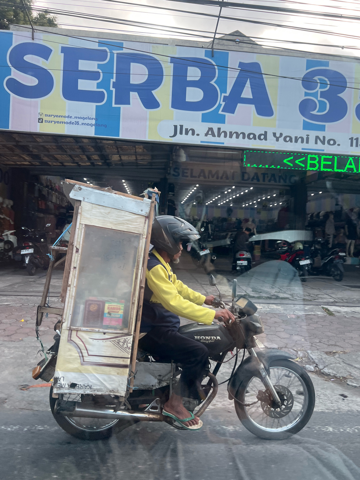 The image shows a man riding a motorcycle with a sidecar in Magelang, Indonesia. The motorcycle is dark-colored and appears to be a Honda. The sidecar is a wooden structure with a glass panel and is decorated with a patterned fabric. Inside the sidecar, there are books and other items. The man is wearing a yellow jacket, black pants, and a helmet. He is also wearing flip-flops. The background shows a storefront with a large banner that reads "SERBA 3". Below the banner, there is a sign with "Jln. Ahmad Yani No. 115" and a green LED display showing "<<BELANJA". There are also other motorcycles and people visible in the background, suggesting a busy street. The overall scene depicts a typical street view in an Indonesian city, with the man and his unique motorcycle-sidecar combination being the central focus.