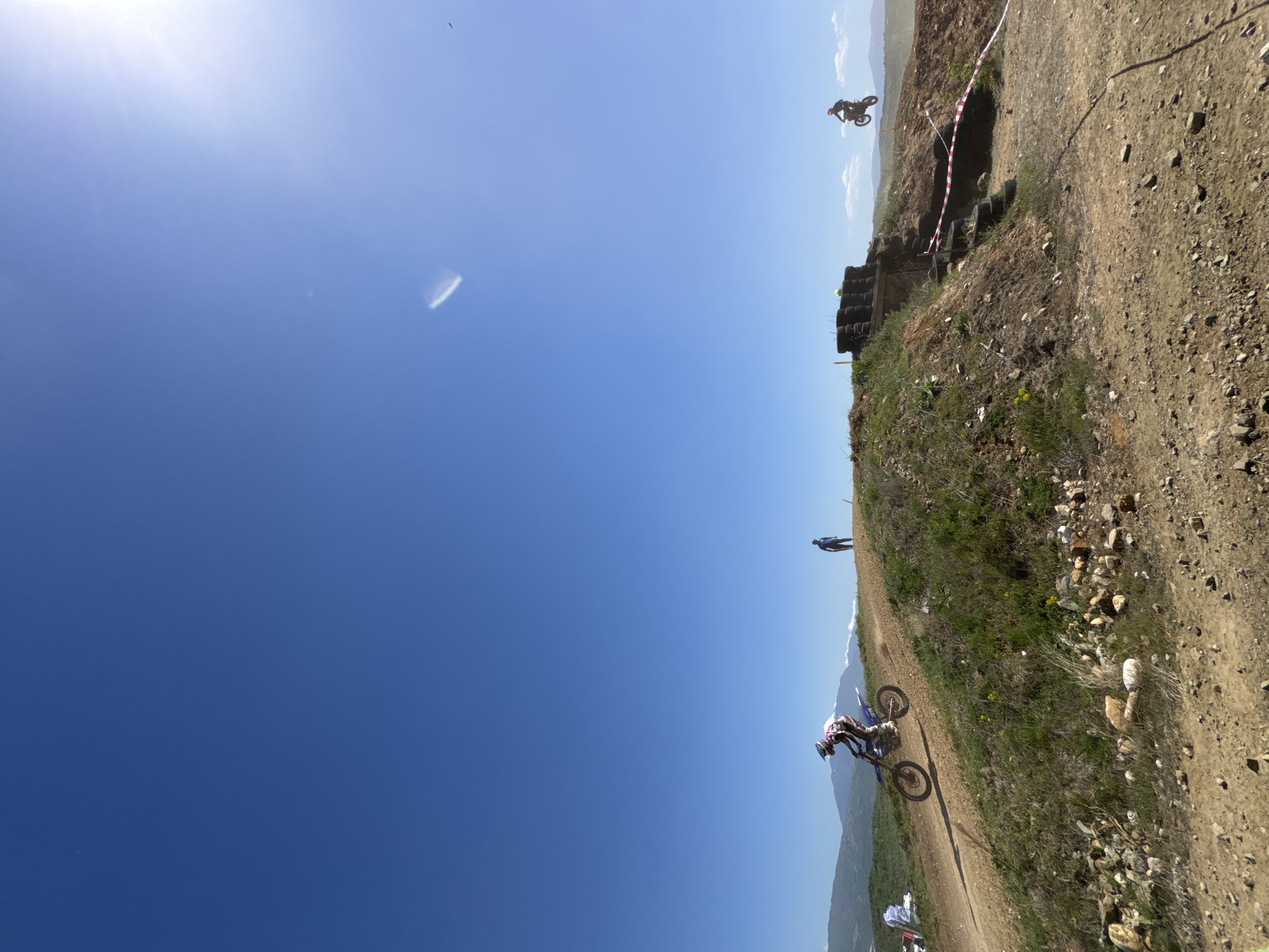 The image depicts a motocross event taking place in Selishte, Bulgaria. A motorcyclist is riding on a dirt track. Another motorcyclist is airborne, having launched off a jump constructed of stacked tires and dirt. Red and white tape demarcates the track. A spectator stands on the hillside. The sky is blue with a few clouds. The landscape is mountainous in the background.