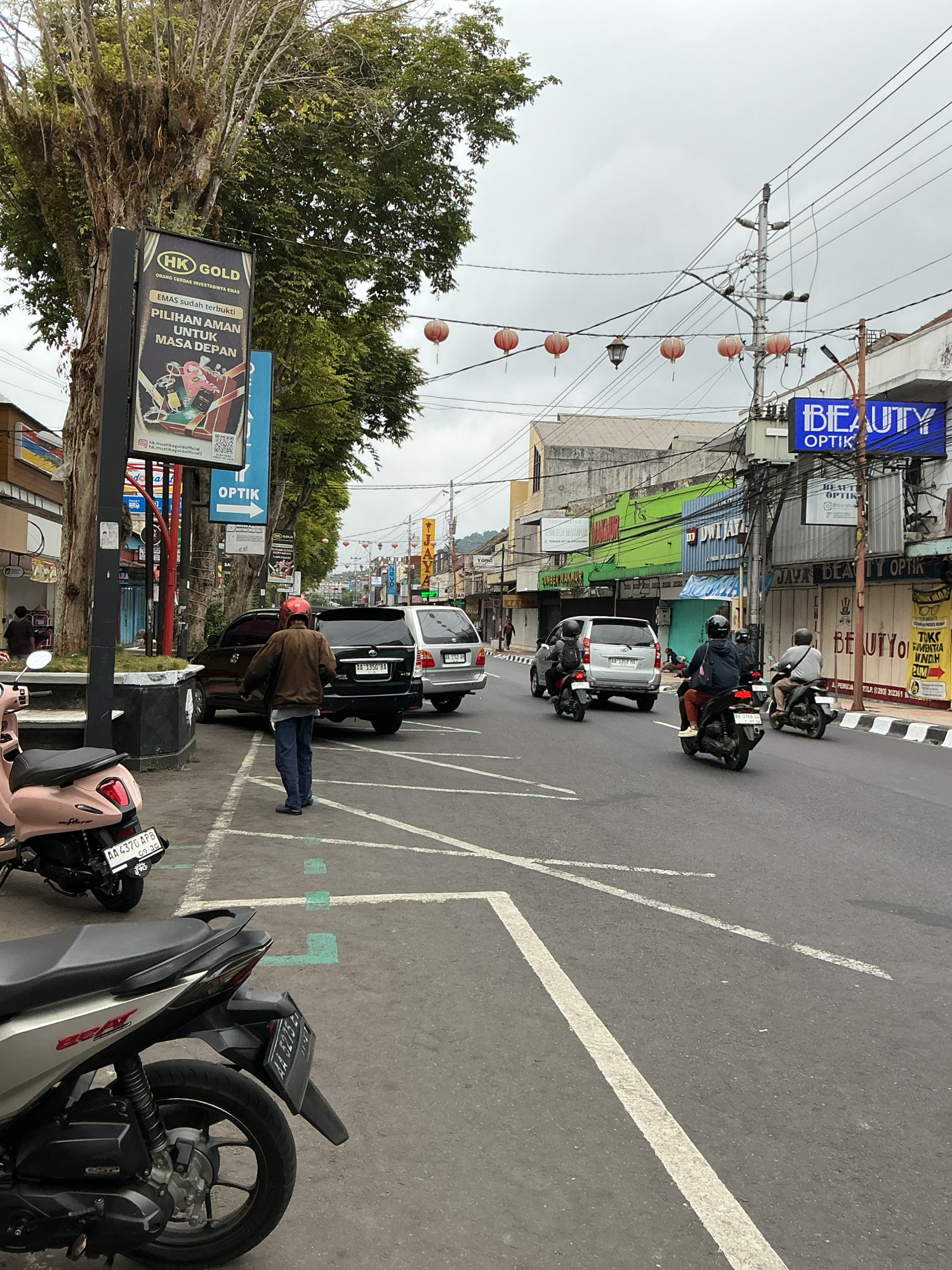 The image captures a street scene in Magelang, Indonesia, with a focus on vehicles and storefronts under an overcast sky. In the foreground, two scooters are parked, one a light pink and the other silver. A person wearing a brown jacket and dark pants, with a red helmet, stands facing away from the camera, looking towards parked cars on the street. Behind them, a dark brown SUV and a silver minivan are parked, their license plates partially visible.
The street is paved with asphalt and marked with white and green parking lines. On the sidewalk to the left, a large tree with a thick trunk stands prominently, with a vertical billboard attached to its base advertising "HK GOLD" and "OPTİK." Further down the street, a row of shops lines the right side, featuring signs like "BEAUTY OPTIK" in blue, and other businesses with colorful awnings and signage in Indonesian. Several red lanterns are strung across the street, suggesting a festive or cultural display.
On the road, three motorcycles are in motion. The closest one has a rider and a passenger, both wearing helmets, heading away from the camera. Another motorcycle further down also carries two individuals. The environment appears to be a commercial area with moderate traffic and pedestrian activity. The overall lighting suggests it is daytime, but the cloudy conditions have softened the light.