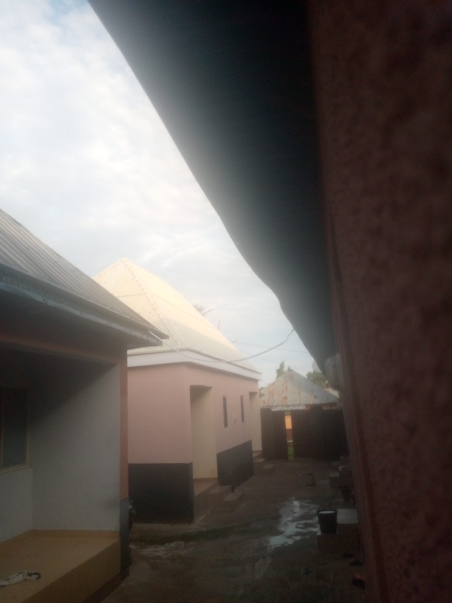 The image captures an outdoor scene in the city of Unknown, Unknown, primarily depicting residential or commercial structures under an overcast sky. A large, out-of-focus, reddish-brown textured surface, likely a wall, occupies the entire right side of the frame, obscuring a significant portion of the scene.
In the midground, on the left, a building with a dark corrugated metal roof and light-colored walls is visible. A gutter runs along its roofline, and a window is present on its left facade. A light-colored raised porch area with a white fabric item rests at its base. To the right of this, a light pink-colored building with dark rectangular sections at its base and narrow vertical apertures is visible. Further in the background, a prominent structure with a large, light-colored, pyramid-shaped corrugated metal roof is centrally located. To the right of this, another building with a dark corrugated metal roof features an open, dark doorway.
The ground area appears wet and reflective, indicative of recent rainfall, with visible puddles and a dark, possibly unpaved, surface. A dark bucket and several light-colored blocks or debris are visible on the wet ground in the lower-right central portion. Sparse green foliage is visible against the light grey, cloudy sky in the upper-middle right. No individuals or animals are present, and no specific actions or interactions are depicted. The overall lighting suggests an overcast day.
