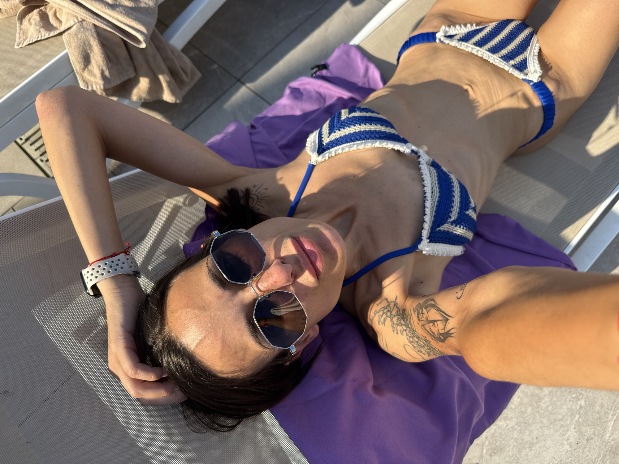 A high-angle, full-body shot captures a woman with dark hair, wearing sunglasses and a blue and white striped bikini, lying on a lounge chair. She is positioned on her back, with her left arm bent at the elbow and her hand resting on her head. The lounge chair appears to be made of a meshed material and is situated outdoors on a tiled surface, possibly a patio or deck. A purple towel is draped over the lounge chair beneath her, and a beige towel is folded on the chair to her right. The woman has tattoos on her left arm. The lighting suggests it is daytime, with sunlight casting shadows. The background is blurred, but a railing and part of a building can be faintly discerned.