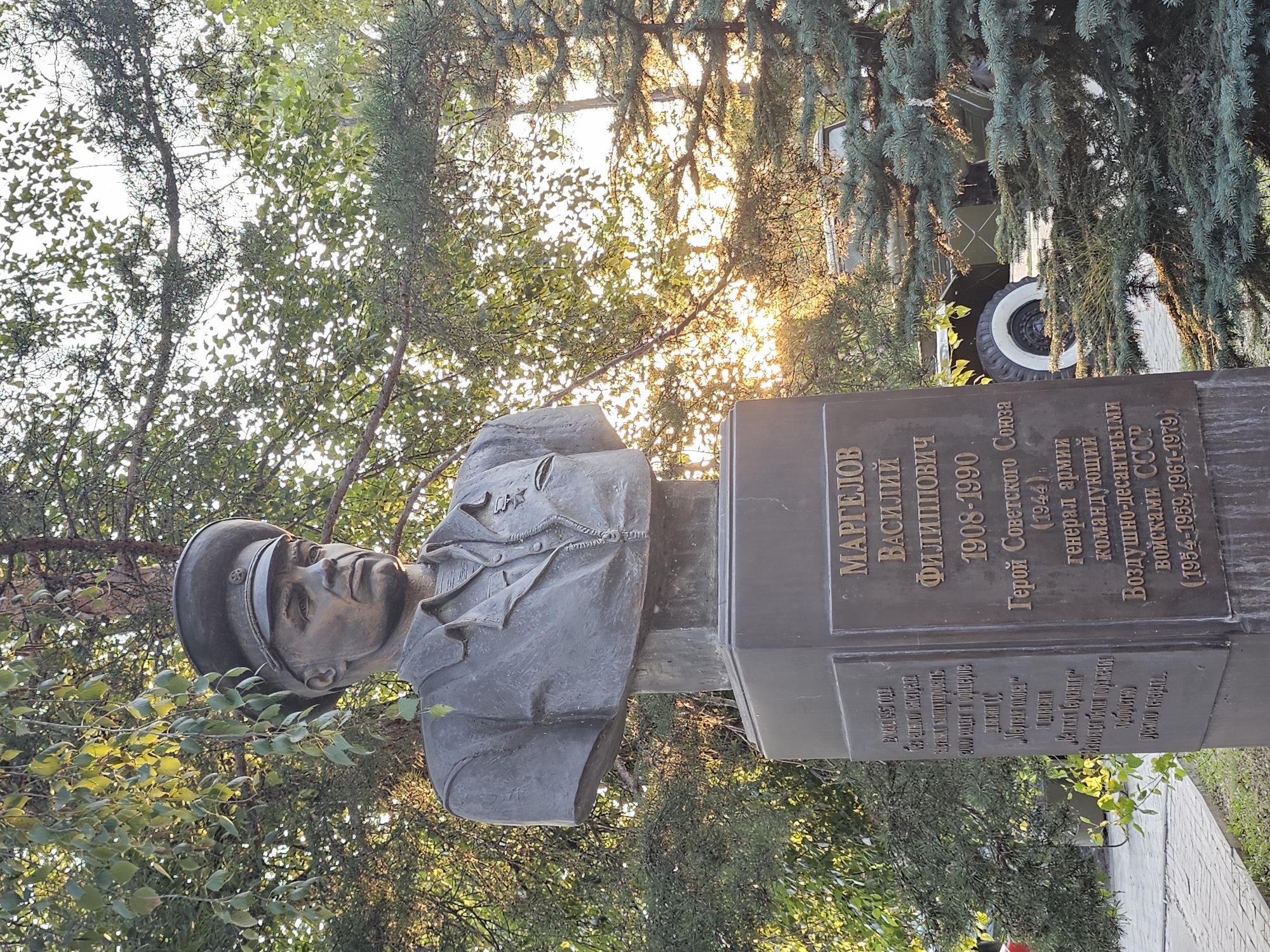 A bust of a Soviet military officer stands on a pedestal in a park-like setting. The bust is made of bronze and depicts the officer in uniform, wearing a cap and a medal on his chest. The pedestal is dark and inscribed with text in Russian. The text on the right side of the pedestal reads: "МАРГЕЛОВ ВАСИЛИЙ ФИЛИППОВИЧ 1908-1990 Герой Советского Союза (1944), генерал армии, командующий Воздушно-десантными войсками СССР (1954-1959, 1961-1979)". This translates to "MARGELOV VASILY PHILIPPOVICH 1908-1990 Hero of the Soviet Union (1944), Army General, Commander of the Airborne Forces of the USSR (1954-1959, 1961-1979)". The bust is located in the city of Cherkessk, Russia, specifically at the Cherkessk Museum of Local Lore.
The background of the image shows lush green trees with sunlight filtering through the leaves, suggesting it is daytime. In the distance, a green military vehicle, possibly a truck or a transport vehicle, is partially visible behind the bust and trees. The ground in front of the pedestal is paved with light-colored interlocking stones. The overall atmosphere is peaceful and commemorative.