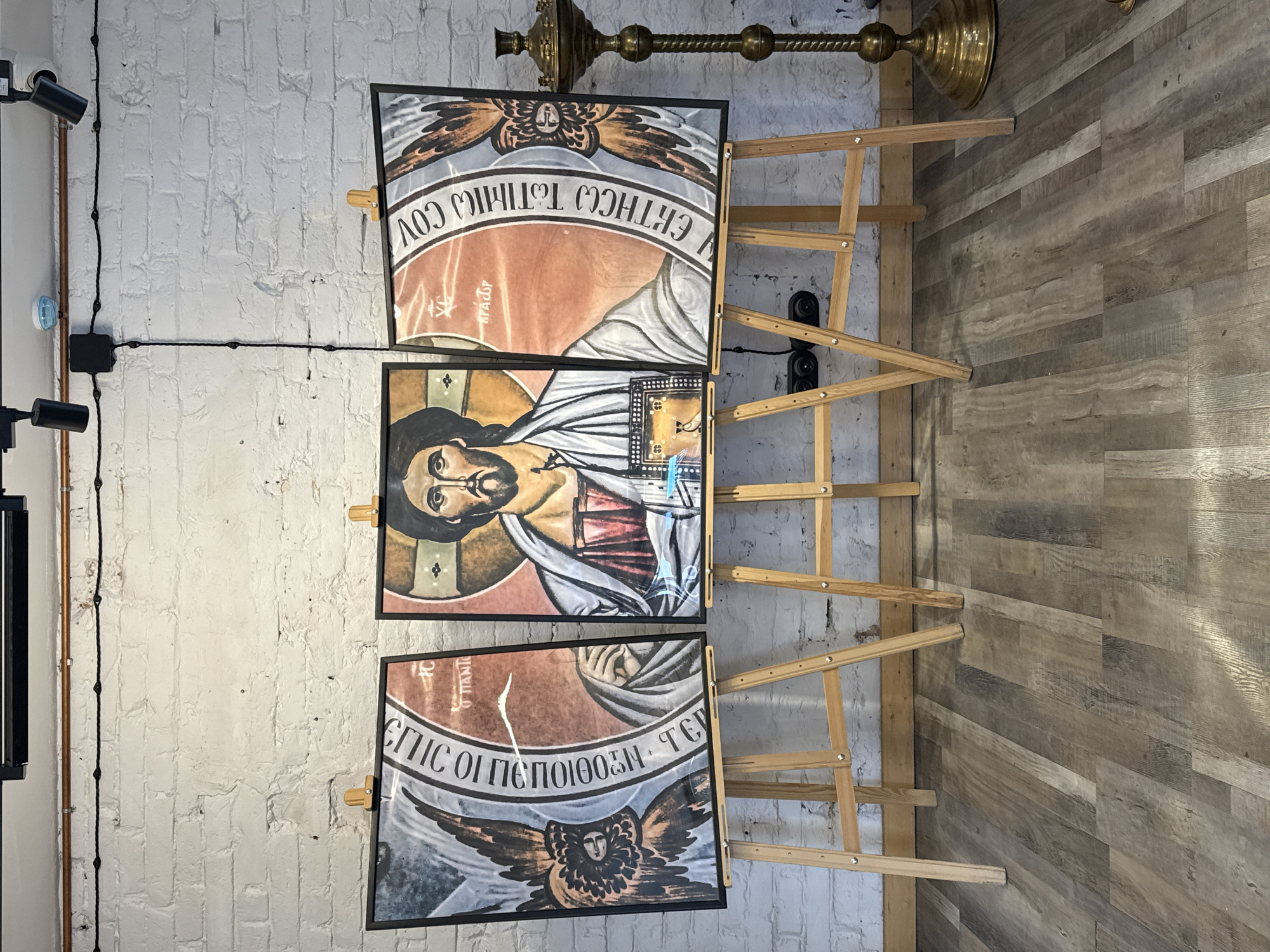 The image shows three religious icons displayed on wooden easels against a white brick wall and a wood-paneled wall. The icons depict religious figures and imagery in a traditional style. The top icon shows an angel or seraph. The middle icon features a portrait of Jesus Christ with a halo. The bottom icon also appears to depict a religious figure with wings, possibly an angel. All three icons are framed in black.
Visible text in the icons is in a script that appears to be Cyrillic or Greek, common in Orthodox Christian iconography. Some readable fragments suggest religious phrases or names. For example, the top icon has text that reads "COV ERTHCC THTHIC COV" and the bottom icon has "EIIC OI IETIOIOOH TCV".
The setting is indoors, with a polished wooden floor and exposed brick. The lighting suggests it is daytime. A brass lamp fixture is visible at the top of the frame. The overall mood is solemn and reverent, consistent with the nature of religious icons. The location context is Yekaterinburg, Russia, which aligns with the likely use of icons in an Orthodox Christian setting.