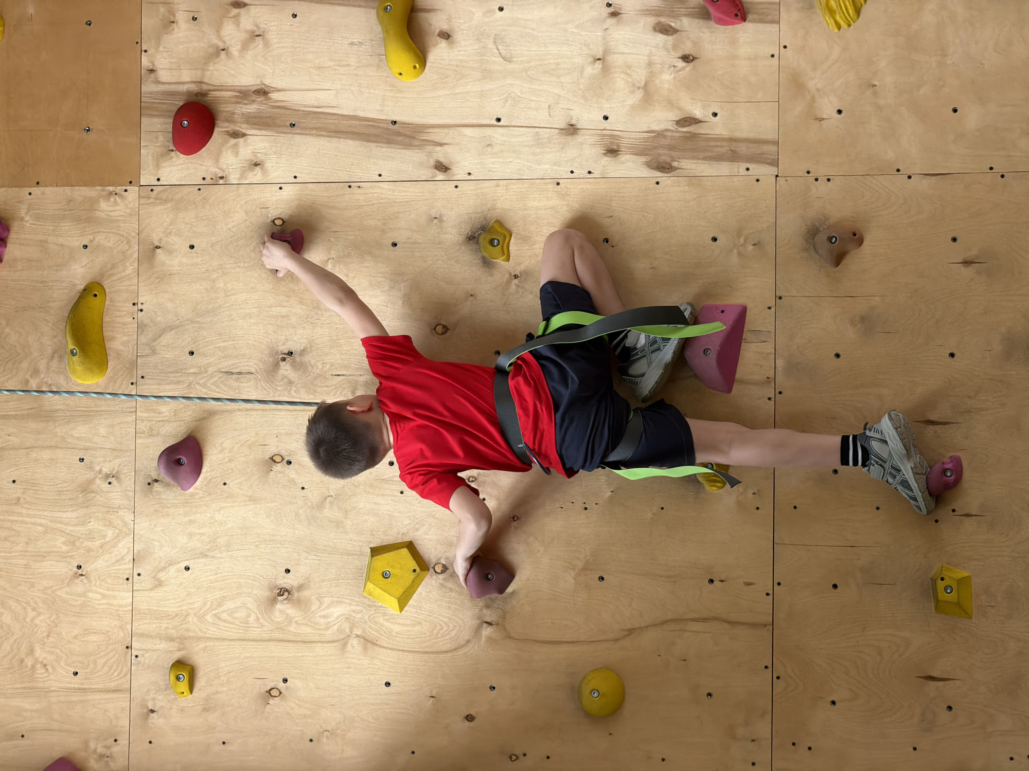 A boy in a red t-shirt, dark shorts, climbing shoes, and a climbing harness is actively climbing an indoor rock climbing wall, likely at an indoor gym or training facility. The wall is constructed of light-colored wood panels with various colored rock climbing holds attached. The boy is positioned horizontally against the wall, using both hands and feet to grip the holds. A climbing rope is visible, suggesting the use of a belaying system. The location is identified as Izhevsk, Russia.