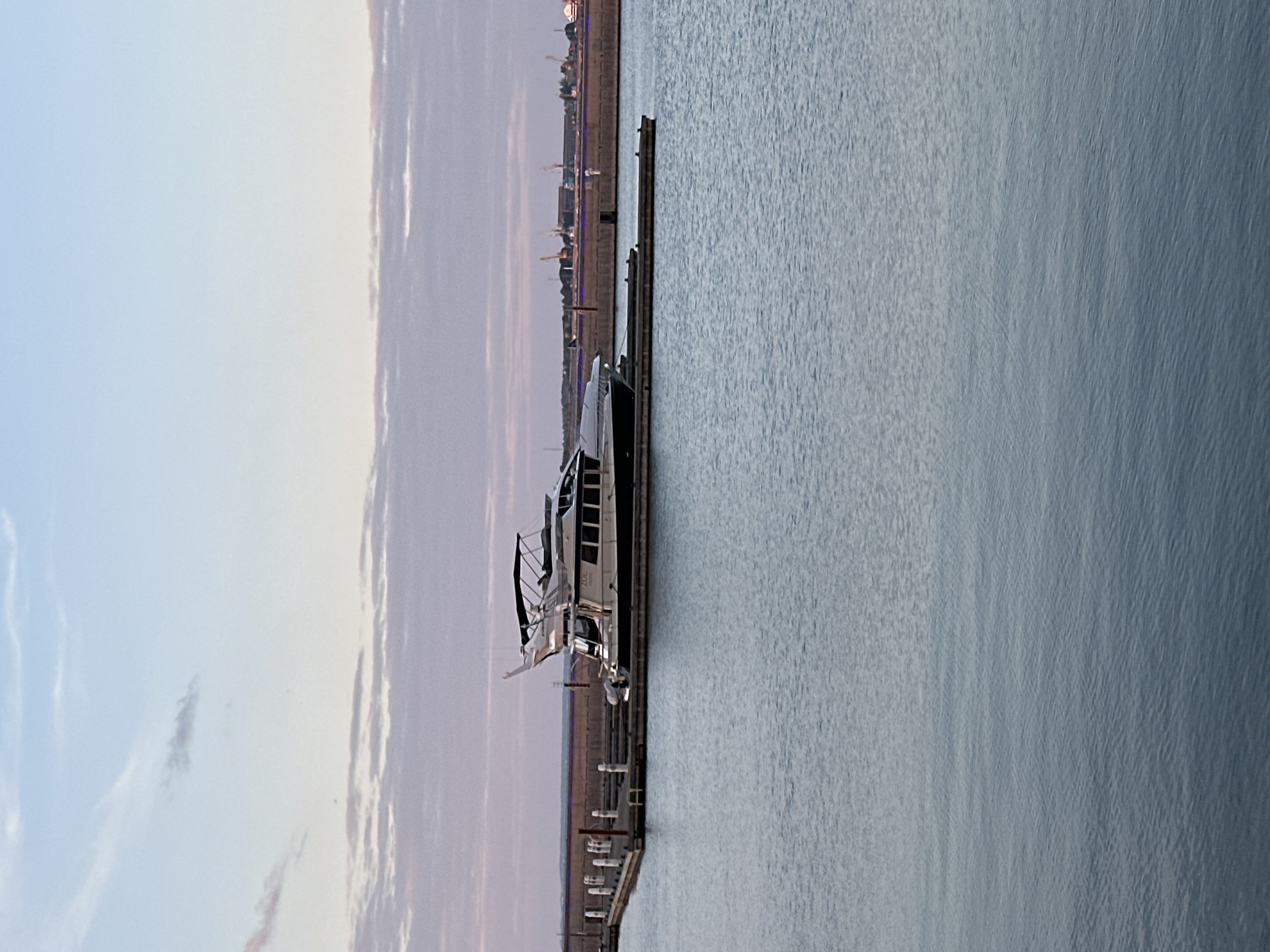 A yacht named "ZOE" is docked at a marina. The yacht is white and black with multiple levels and a flybridge. It is moored to a wooden pier with white bollards. The marina is situated on a body of water that appears calm. In the background, there is a long, concrete seawall or breakwater. Beyond the seawall, a cityscape with industrial elements and cranes is visible, suggesting an urban or port environment. The sky above is a pale blue with scattered wispy clouds, indicating either early morning or late evening light. The overall scene depicts a peaceful harbor setting. The location is not identifiable as Tallinn, Estonia, based on visual cues alone.
