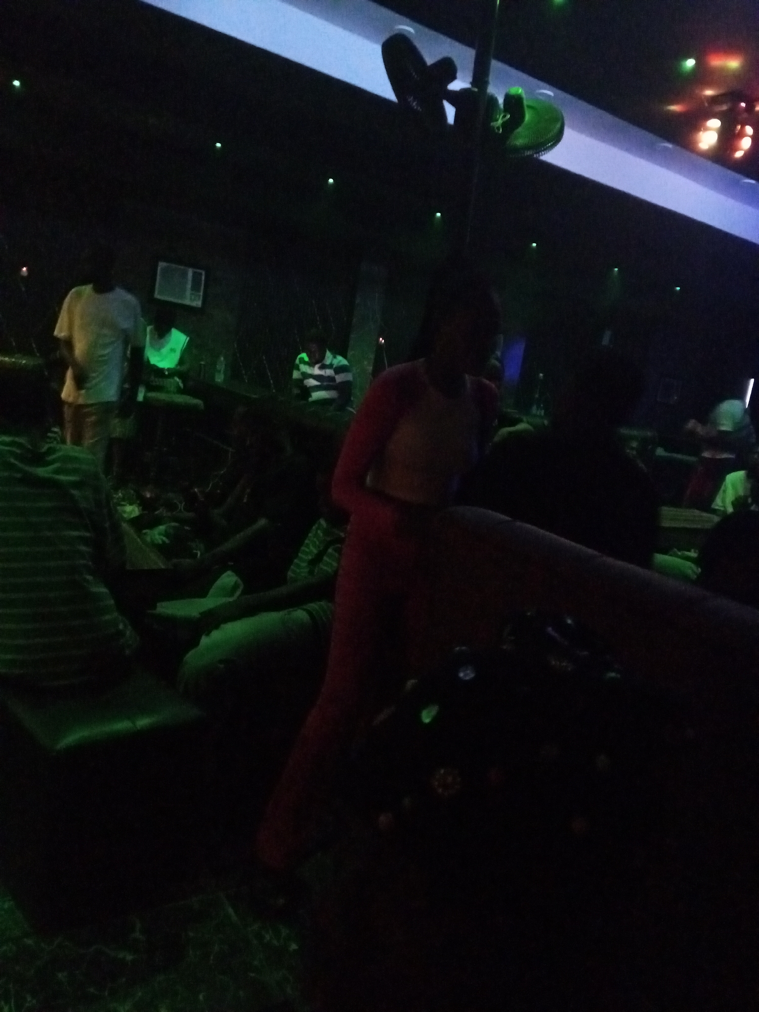 The image appears to be taken in a dimly lit indoor space, potentially a nightclub or bar, as suggested by the green lighting and the presence of people seated and standing. There are multiple individuals in the scene, including a person wearing a white t-shirt, another person in a neon green vest or shirt, and a person in red clothing towards the foreground. The setting contains seating areas with cushions, and a ceiling fan is visible in the upper portion of the frame. Overall, the environment seems crowded and dimly lit, indicative of a social gathering in an entertainment venue. The location of the image within the city of Unknown, Unknown is not possible to determine given available information.