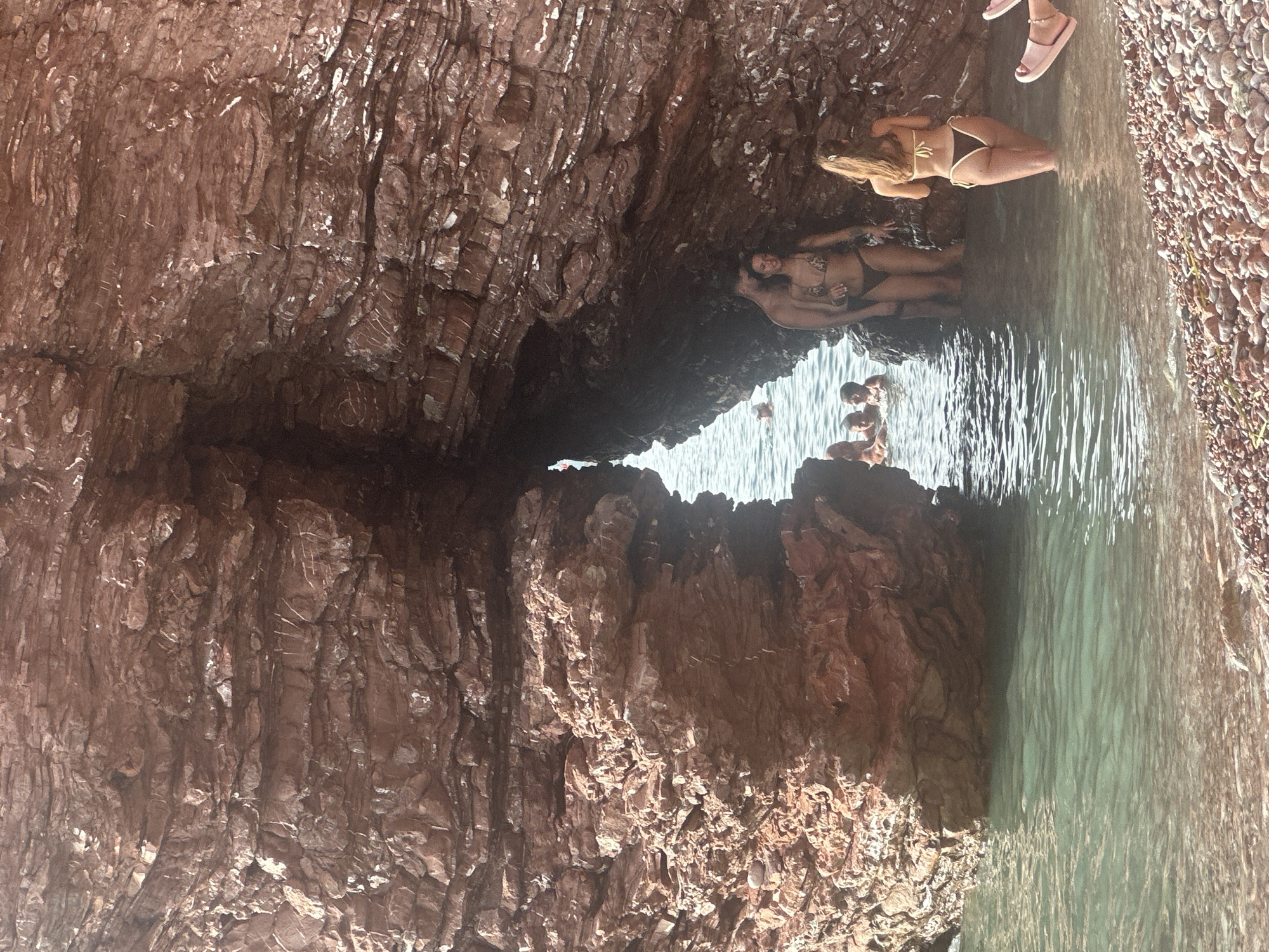 A group of people are gathered in and around a natural rock archway at a beach, possibly located in Sveti Stefan, Montenegro. The rock formation is reddish-brown and appears layered, with rough textures and some lighter mineral deposits. The water is clear and turquoise, with small pebbles visible on the seabed and along the shore.
In the foreground, on the right, a person is wearing pink flip-flops. Slightly to the left, a woman in a yellow bikini stands in the shallow water, facing away from the camera. Behind her, partially obscured by the rock, a couple stands close together. The man is wearing dark shorts and the woman is in a leopard print bikini. To the left of this couple, other individuals are visible in the water. In the background, through the archway, more people are swimming or standing in the water. The sun is shining, creating bright light and some shadows.