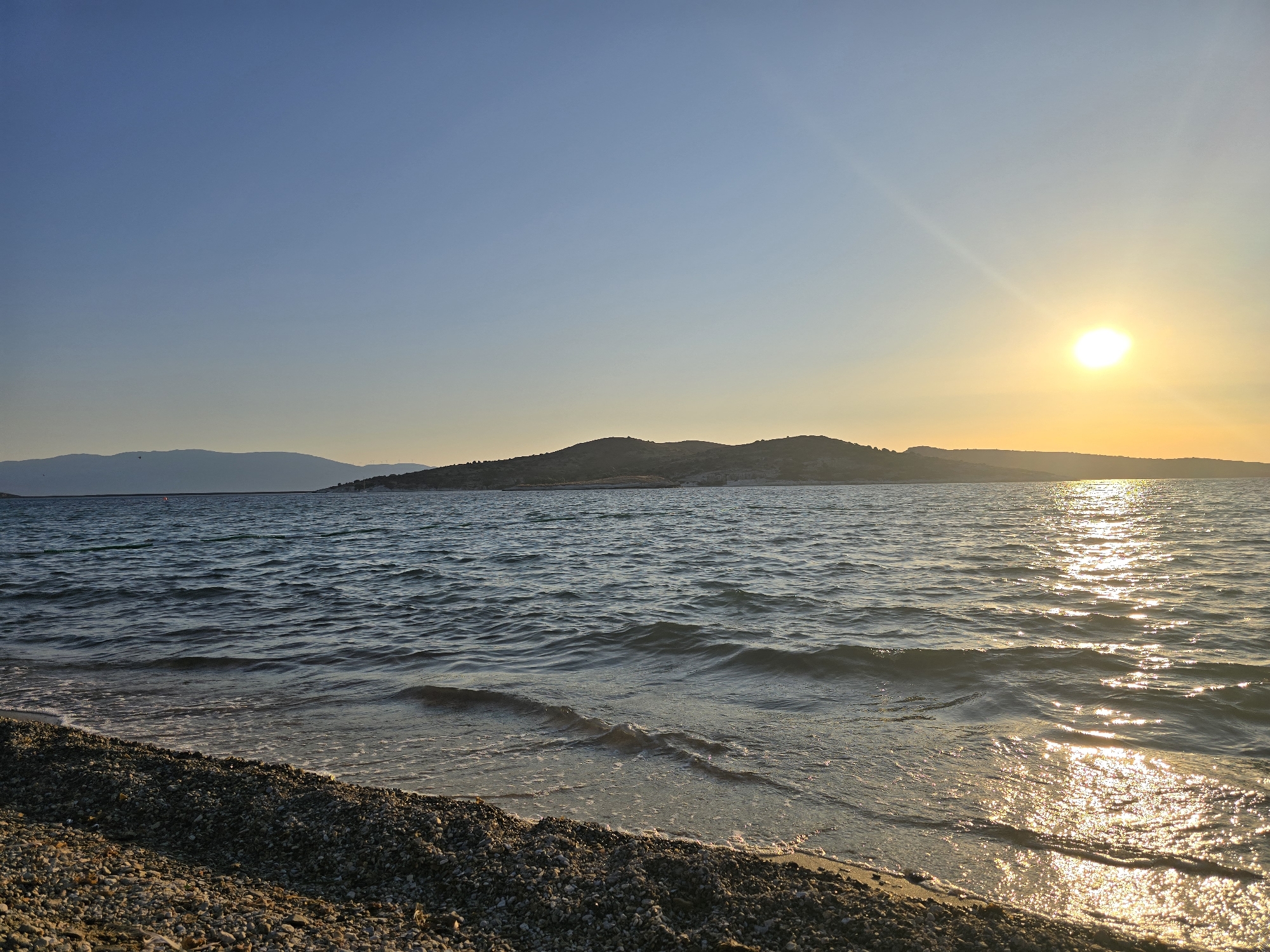 A serene wide-angle photograph captures a pebbled beach leading into the calm Aegean Sea at sunset in Foça, Turkey. The sun, a bright orb near the horizon, casts a golden shimmer across the water, with gentle waves lapping the shore. In the distance, a hilly, tree-lined coastline is silhouetted against the gradient sky, transitioning from a clear azure blue at the top to a soft yellow and orange hue near the horizon. The beach itself is covered in small, dark gray and brown pebbles. The lighting suggests late afternoon or early evening, creating a tranquil and picturesque coastal scene. No people are visible in the frame.