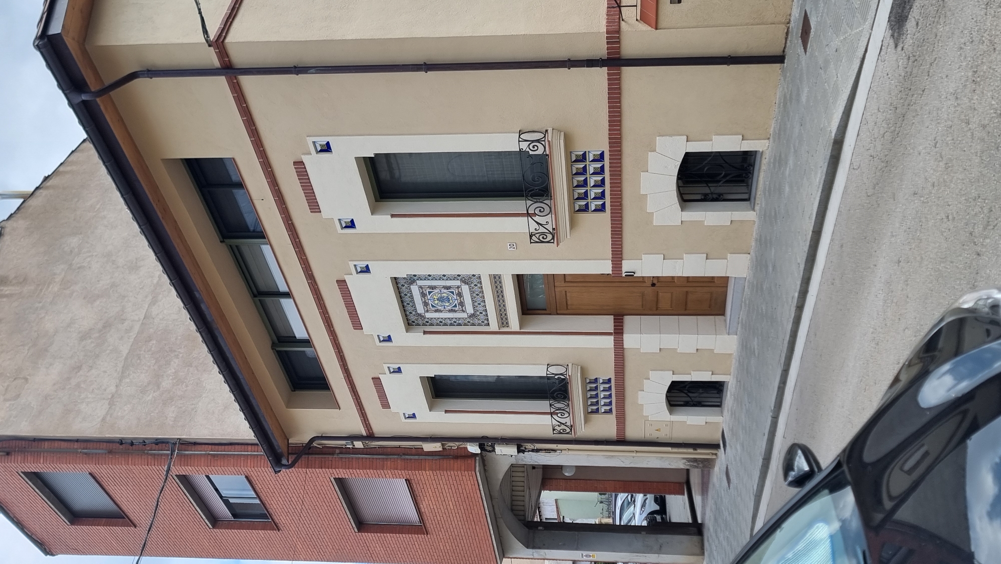 A beige building with decorative blue and white tiles and dark brown trim is viewed from the street in Olot, Spain. The building features a wooden door, several tall windows with ornate black railings, and arched basement windows with black grates. To the left of the beige building, a red brick building with multiple windows and grey shutters rises higher. The street in front of the buildings is paved with grey stones, and a black car is partially visible in the foreground. The sky is overcast.