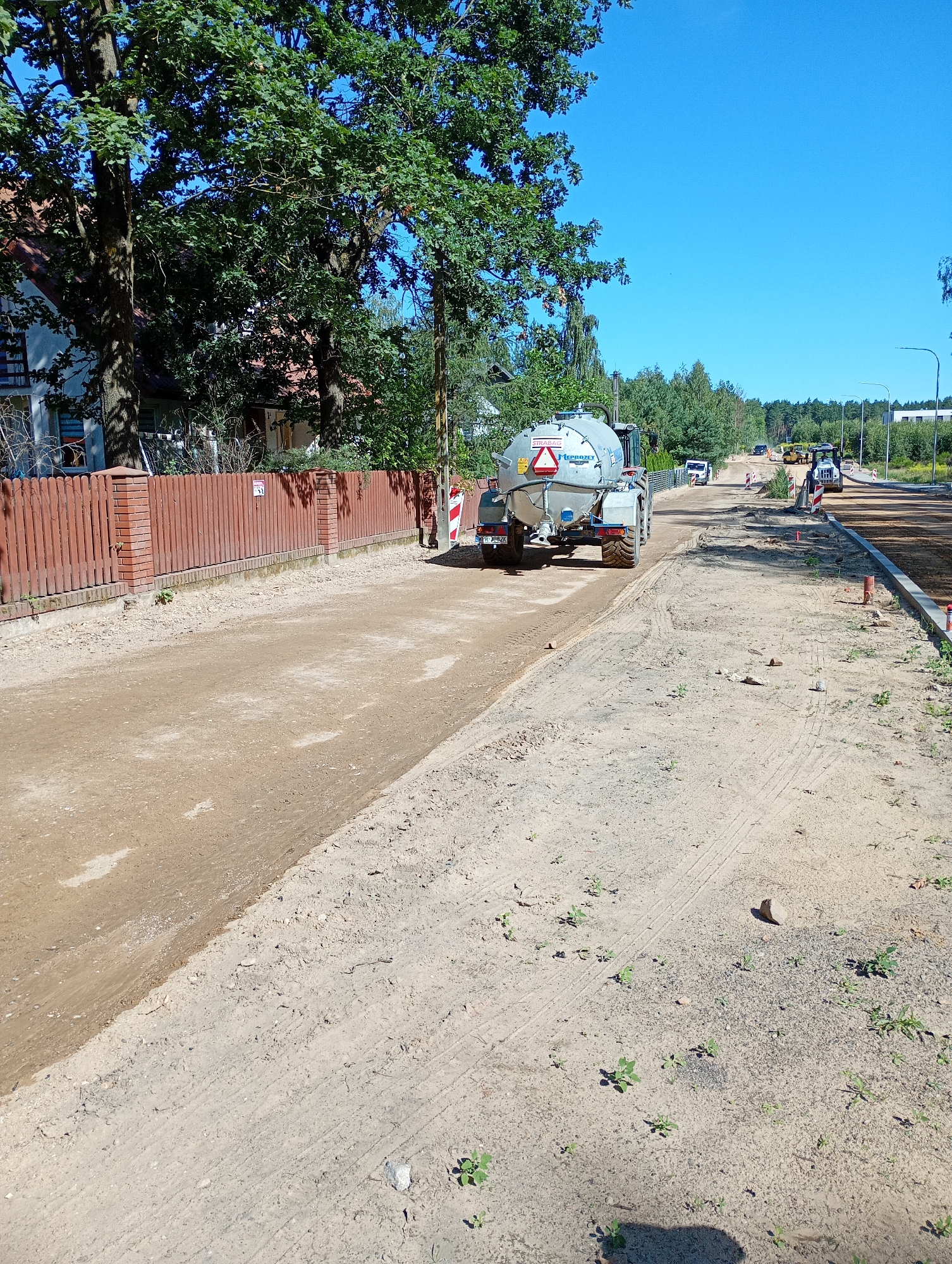 This is a photograph taken on a sunny day in Grabówka, Poland, depicting a construction site. The foreground shows a dirt road under construction, with tire tracks from heavy machinery visible. A red wooden fence with brick pillars lines the left side of the road, separating it from a property with a house and large trees.
In the middle ground, a large, gray water truck with the logo "STRABAG" is positioned on the dirt road, appearing to be part of the construction process. Further down the road, other construction vehicles, including a yellow road roller and what appears to be a backhoe, are visible. The road itself is unpaved and appears to be in the early stages of development, with curbs and some pavement visible in the distance.
The background consists of a clear blue sky and a line of trees, suggesting a semi-rural or suburban environment. The overall scene captures a moment in the ongoing development and infrastructure work within the city of Grabówka.