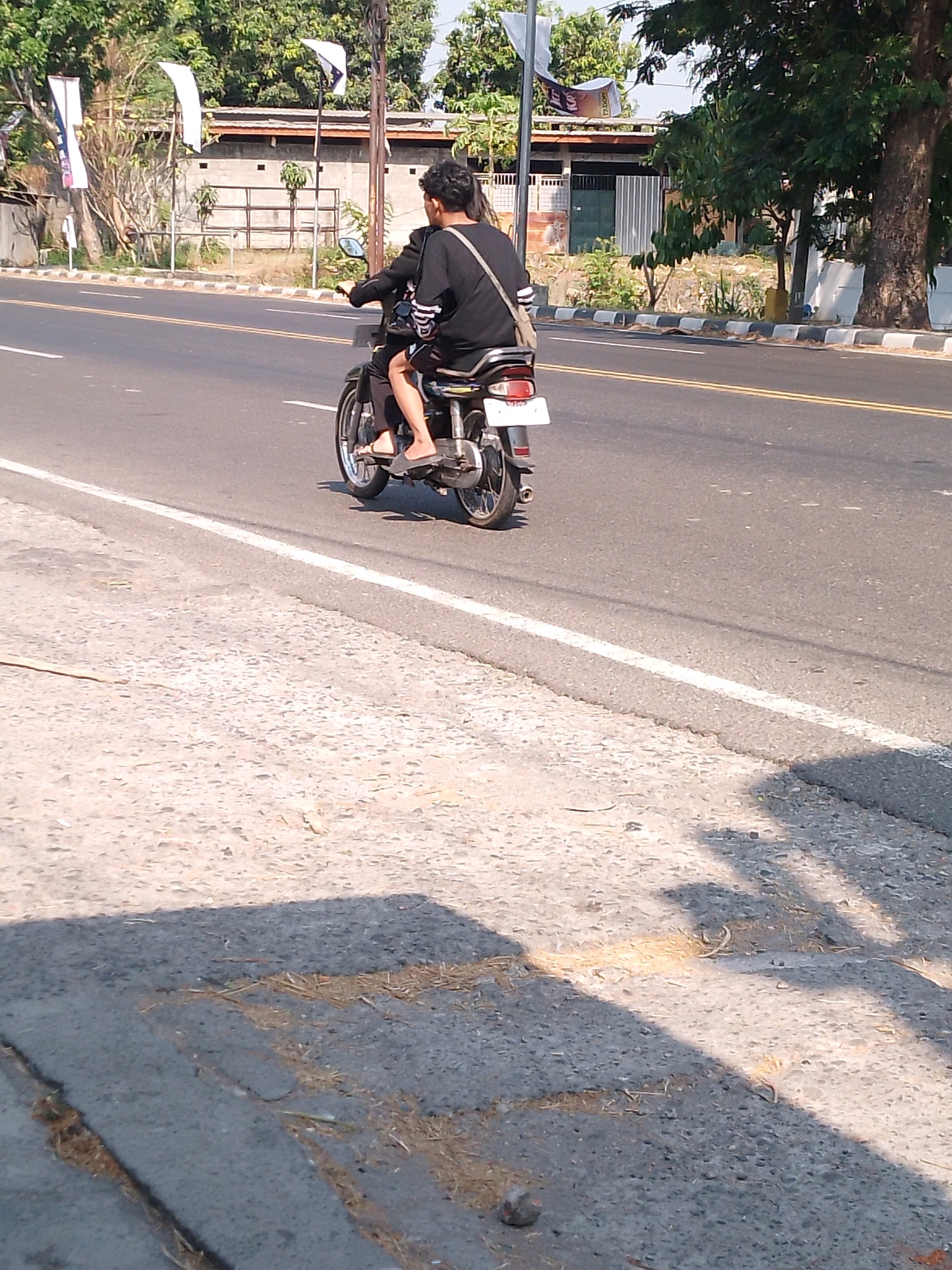 A medium shot captures a person riding a motorcycle on a paved road in Kutorejo, Indonesia. The person, viewed from behind, wears a black shirt and shorts, with a bag slung over their shoulder. The motorcycle has a white license plate. The road is marked with double yellow lines and a white dividing line. A concrete sidewalk with some debris lines the road. In the background, there are buildings, trees, and flags. The scene is lit by sunlight, casting shadows on the road and sidewalk.
