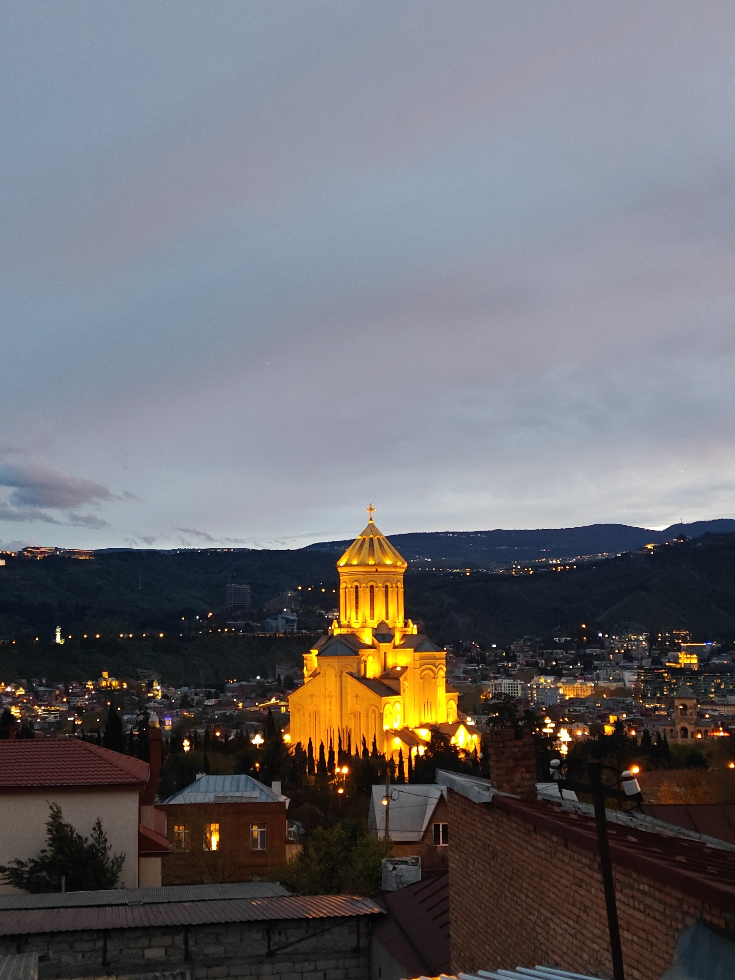 This photograph captures a twilight panoramic view of Tbilisi, Georgia, centered on the magnificent Holy Trinity Cathedral (Sameba Cathedral) brightly illuminated on a hilltop. The sky is a blend of soft blue, purple, and gray hues, indicative of dusk or dawn, with visible clouds.
The Holy Trinity Cathedral stands out prominently, bathed in a warm, golden-orange glow that emphasizes its grand architecture, including its large central golden dome, smaller domes, and detailed facades with arched windows. Dark, slender trees are visible around the base of the cathedral.
Beyond the illuminated cathedral, dark hills and mountains form the background, stretching across the horizon. These hills are dotted with numerous small, twinkling lights, suggesting further city development or infrastructure. The broader cityscape below and around the cathedral also shines with countless artificial lights, creating a vibrant evening atmosphere.
In the immediate foreground, several residential rooftops are visible, featuring red tiles, corrugated metal, and brick walls. Some windows in these foreground buildings emit a warm light, adding a touch of domesticity to the grand scene. The perspective suggests the image was captured from an elevated position, looking over local rooftops towards the city's iconic landmark.
The overall scene conveys a sense of tranquility and grandeur as the city transitions from day to night. There are no people visible, and no specific event is taking place. The weather appears calm, with no indication of precipitation. No text is visible in the image.