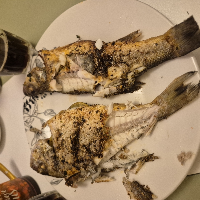Two cooked fish are centered on a white plate. The fish are split open, revealing their white flesh and bones. Their skin is golden brown and speckled with dark brown markings, possibly from spices or charring. The tails of the fish are intact and extend towards the top and bottom right of the frame. A dark glass bottle sits to the left, partially obscured, and a brown jar is visible at the bottom left corner. The plate rests on a light-colored surface.