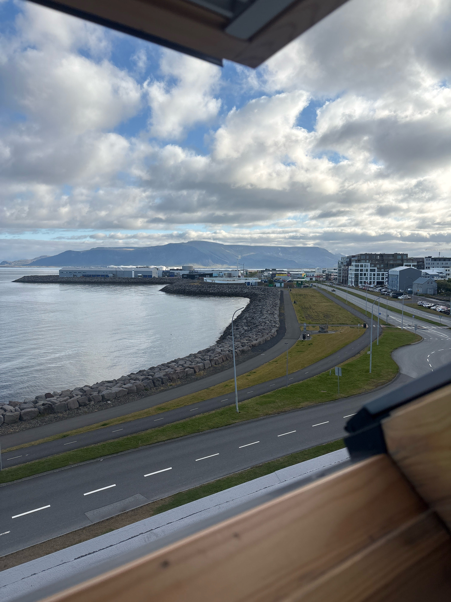 This is an outdoor, eye-level, medium shot of a coastal road in Reykjavik, Iceland. The image features a road that curves along the coastline, with a breakwater made of large stones separating it from the water. Buildings, including modern apartment complexes and industrial structures, are visible along the road. The sea is calm and reflects the cloudy sky, which is dotted with white clouds against a light blue background. A distant mountain range is visible on the horizon. The foreground of the image is partially obscured by the edge of a wooden structure, possibly a balcony or window frame. There are no people visible in the scene, and no specific actions or interactions are taking place. The environment appears to be a developed urban area situated by the sea.