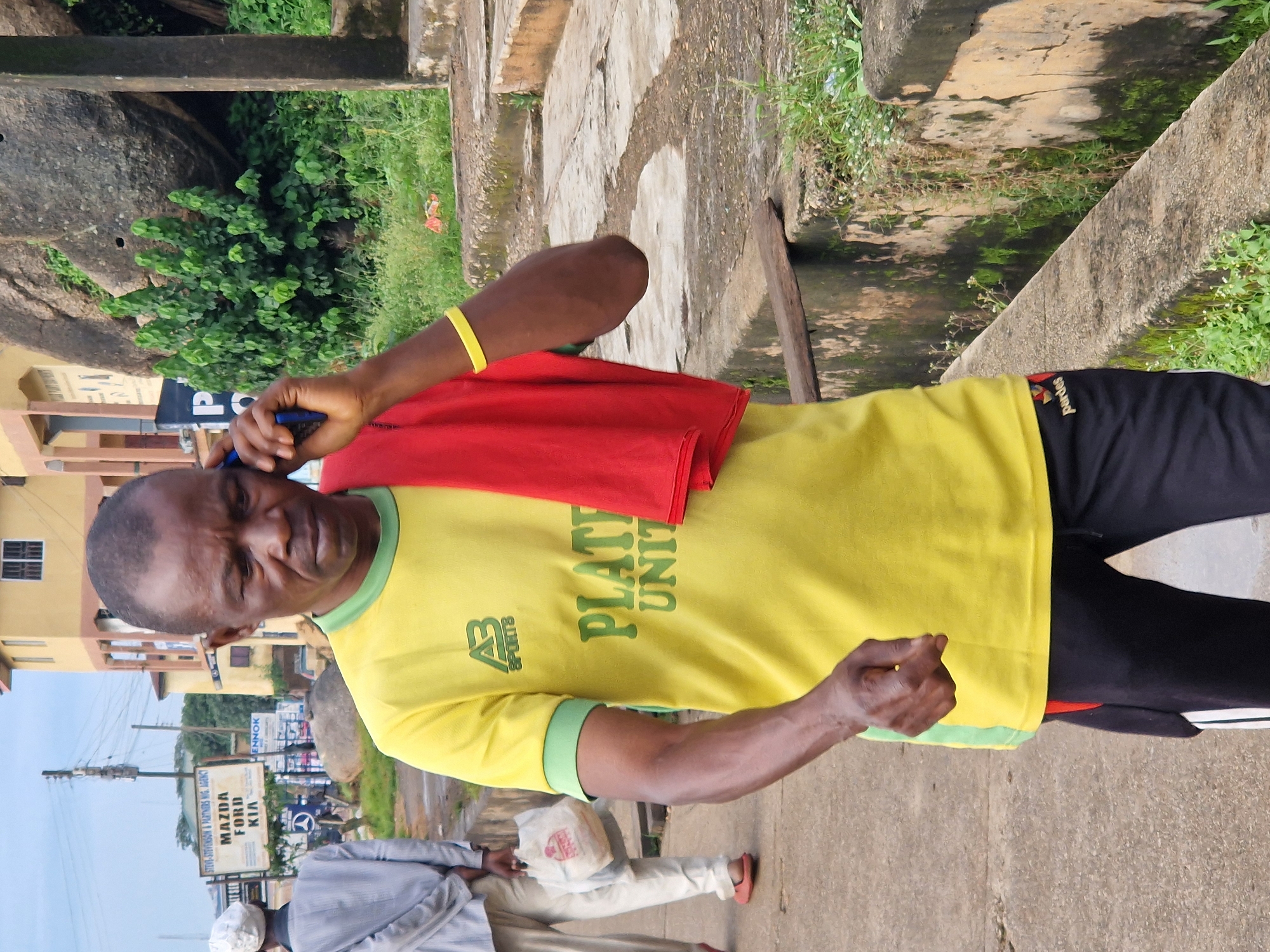 A Black man, appearing to be middle-aged, walks towards the viewer and talks on a mobile phone. He is wearing a yellow t-shirt with green trim and the words "PLATEAU UNITED" in green, black athletic pants with a red stripe, and a red cloth draped over his right shoulder. A yellow bracelet is on his left wrist. He is on a paved sidewalk next to a low, weathered wall. In the background, a second Black man, wearing a white cap and light-colored clothing, walks away from the viewer carrying a white plastic bag. A signpost with "STEVE-STEVENSON & PARTNERS NIG. AGENCY MAZDA FORD KIA" is visible behind him. Further in the background are buildings, a large rock formation, and lush green vegetation. The scene appears to be a street in New Stadium, Nigeria.