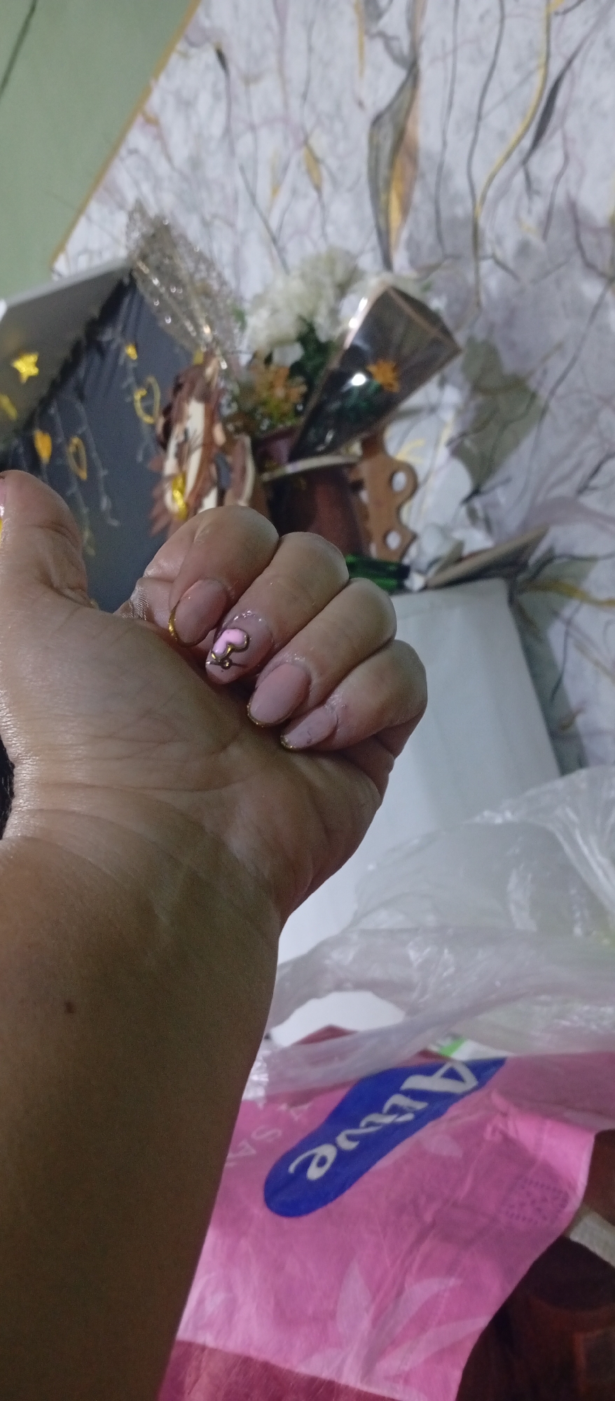 A person's hand is displayed with pink manicured nails, one adorned with a pink heart design. The background includes decorative items such as flowers and a decorative box. The lower right corner contains a pink package labeled with the word "Alive." The exact location within the city of Juan Griego, Venezuela, cannot be determined from this image.