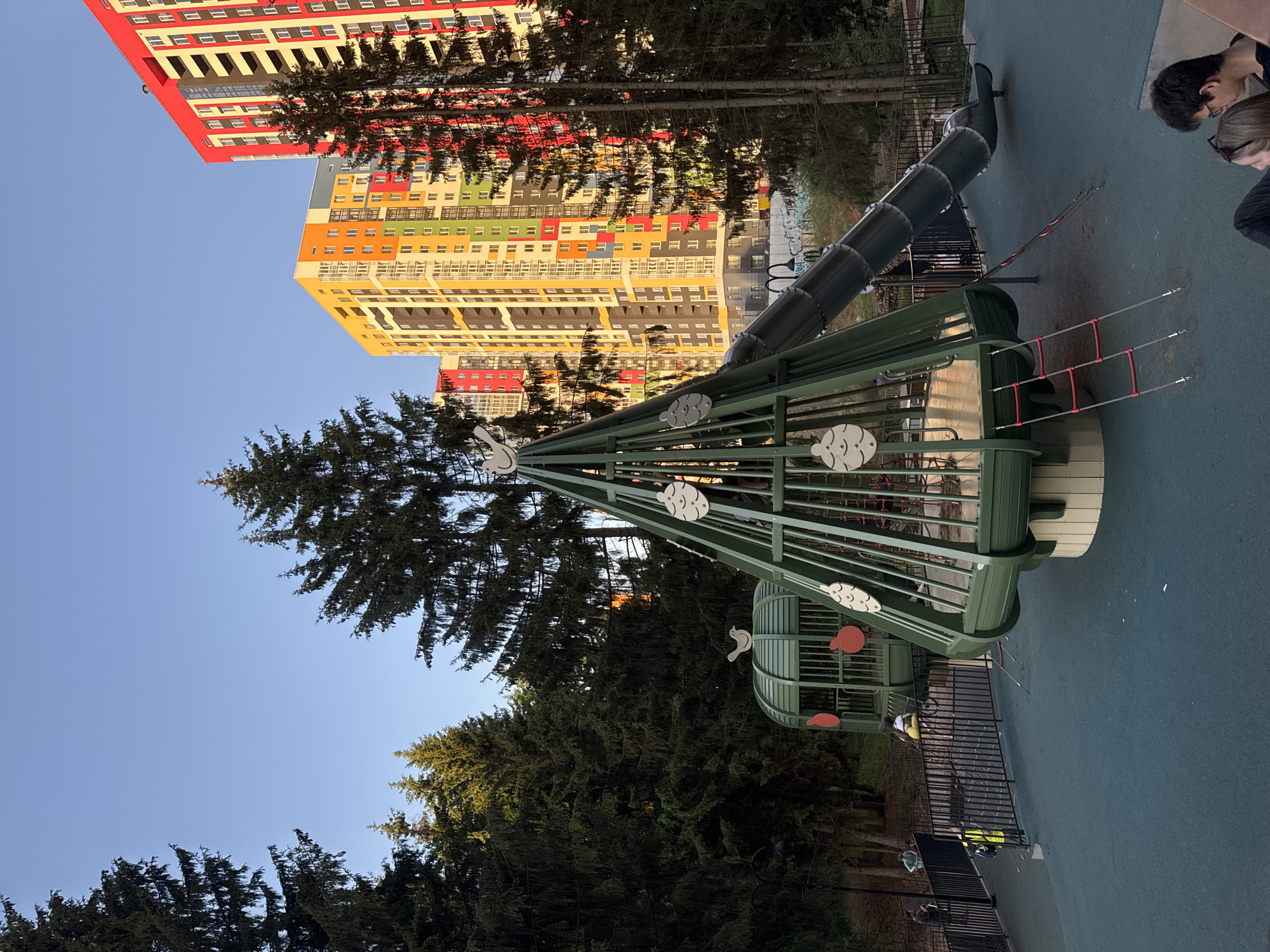 The image shows a playground with modern play equipment in front of a tall, colorful apartment building. The playground features a green climbing structure with cloud and bird decorations and a tunnel slide. Trees surround the playground. The location is Izhevsk, Russia, based on the architectural style of the apartment building. Two people are partially visible in the upper right corner of the image.