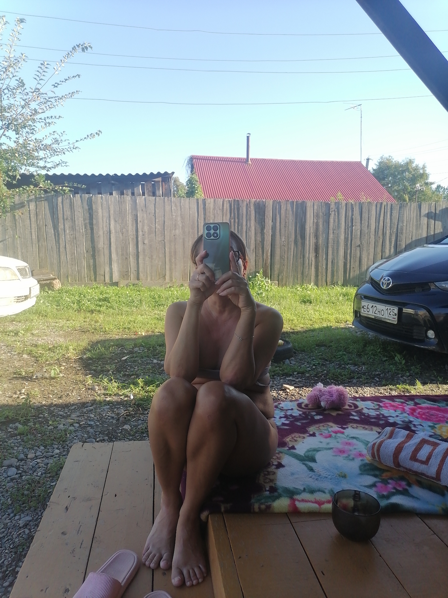 A medium shot captures a woman sitting outdoors on a wooden deck, holding up a smartphone to take a selfie. She is positioned on the right side of the frame, with her legs crossed and bent. The woman appears to be wearing a dark bikini top and bottoms. Her face is obscured by the phone, but she has on glasses and a bracelet on her left wrist.
In front of her, on a colorful patterned blanket, is a small fluffy pink object, a rolled-up towel with orange and white stripes, and a dark bowl. To the left of the woman, a pair of pink slides are visible on the deck.
The background reveals a weathered wooden fence, a strip of green grass, and some gravel. Beyond the fence, a house with a red corrugated roof is visible. To the left of the fence, a white car is parked. To the right of the fence, a black Toyota is parked. The sky is clear and blue, with power lines running across the top of the frame. The scene appears to be in a rural setting. The license plate on the black car reads "E6 12HO 125", indicating it is from Russia. The presence of the Russian license plate and the visible architectural style suggests the location is likely in Novopokrovka, Russia.