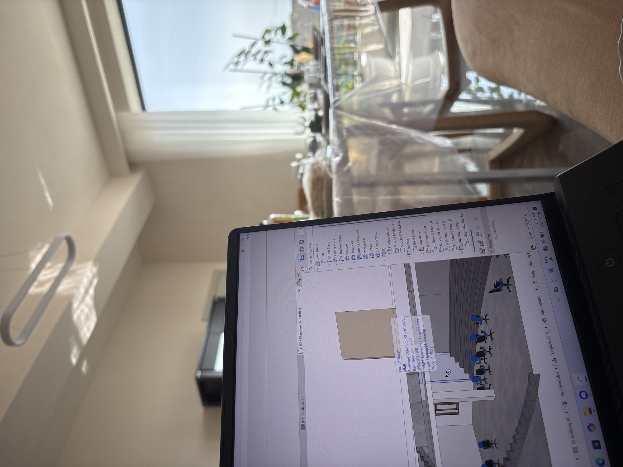 The image shows a laptop screen displaying architectural design software, possibly ArchiCAD, with a 3D model of a building interior. The design appears to show a multi-level space with rows of chairs, potentially an auditorium or lecture hall. On the left side of the laptop, the camera captures a room with a window revealing an outdoor view, possibly through a window or balcony. The presence of indoor plants on a table or shelf suggests a domestic or office environment.
Based on the laptop screen capture, one can infer the user is working on architectural plans for a building that may feature a lecture hall with multiple tiers and rows of seating. The laptop also displays text in Armenian, specifically "Արտաքին պատեր" which translates to "Exterior Walls."
The location of Abovyan, Armenia cannot be determined from the image alone. The architecture visible on the screen does not provide sufficient context.
