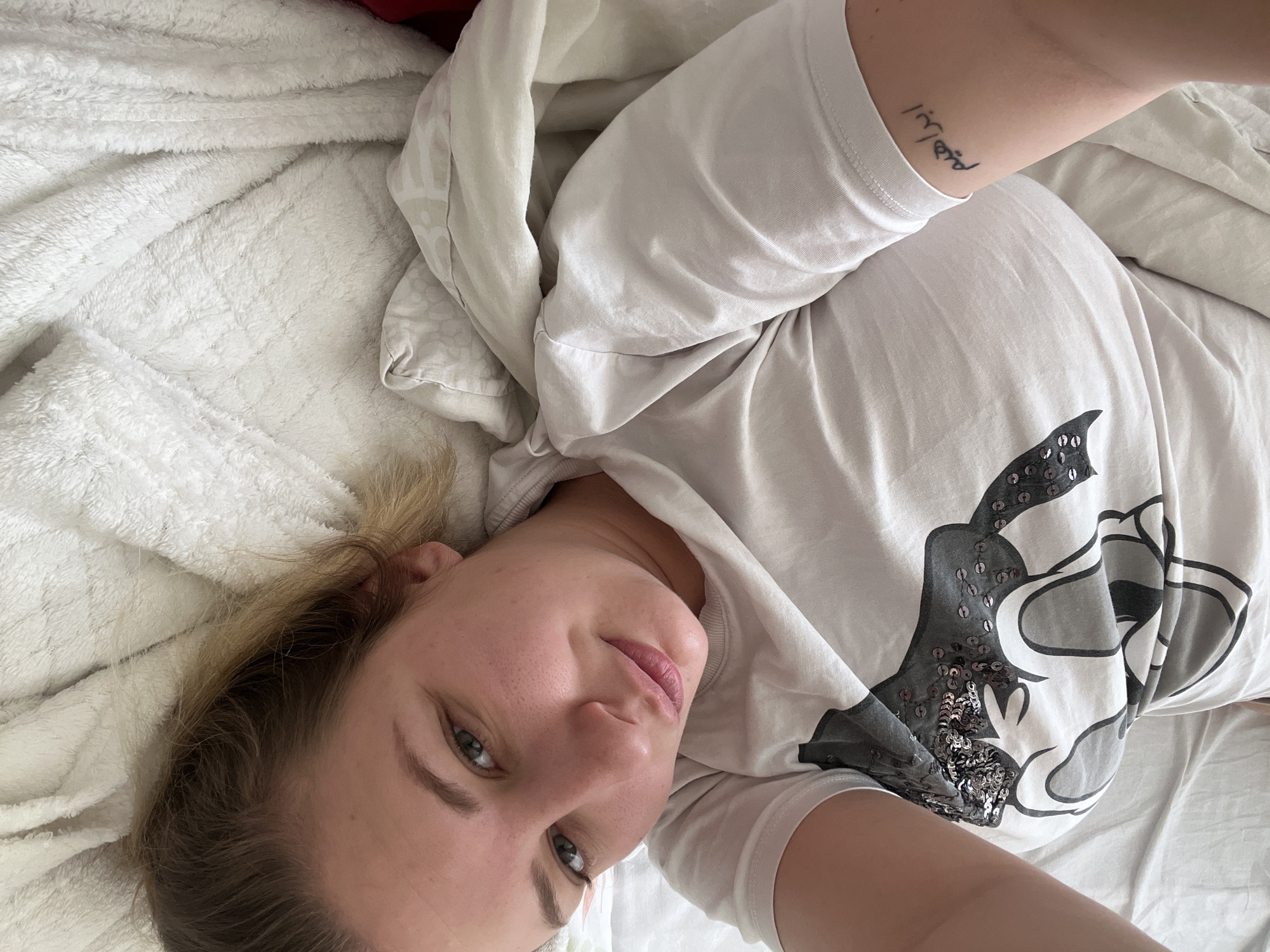A woman with blonde hair and light skin is lying on a white bed. She is wearing a white t-shirt with a Mickey Mouse design that has sequins on it. The woman has a tattoo on her arm. The background consists of white bedding. The location is unknown.