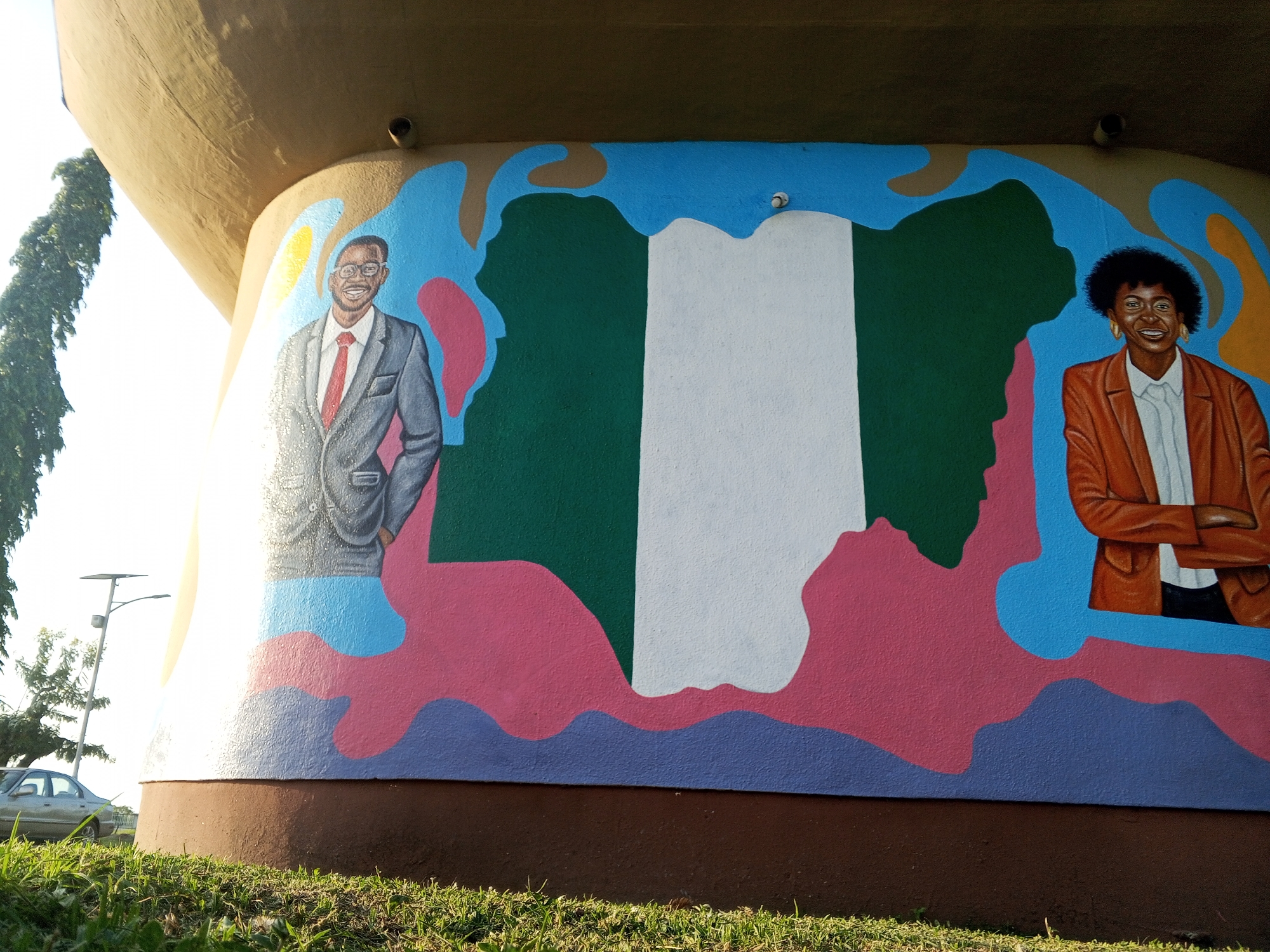 The image shows a mural painted on a rounded, brown-based structure, likely a water tower, located in Tawpo, Nigeria. The mural features a stylized representation of the Nigerian flag, with green, white, and green vertical stripes. Flanking the flag are two portraits of smiling individuals: a man in a grey suit with a red tie to the left, and a woman in a brown blazer to the right. The background surrounding the flag and portraits is a colorful abstract pattern of blue, pink, and brown. The structure sits on a grassy area. A tree and street light pole are visible in the background. A car is parked nearby.
