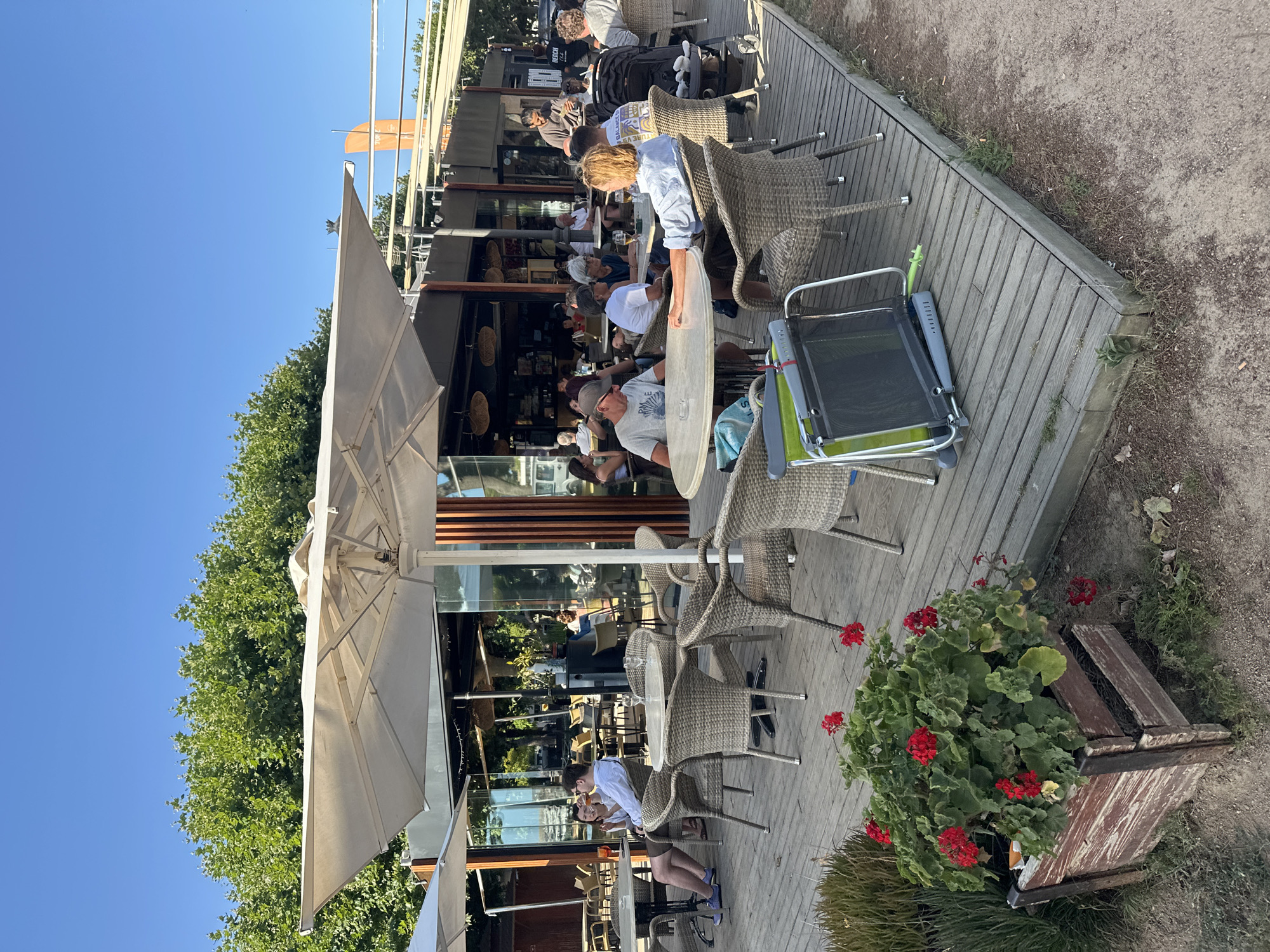 The image depicts an outdoor seating area of a beachfront restaurant in Sant Feliu De Guíxols, Spain, with a clear blue sky overhead and lush green trees in the background. The foreground features a wooden deck furnished with wicker chairs and tables, some occupied by patrons enjoying drinks and conversation. A planter box with red geraniums sits on the left side of the deck. In the middle ground, the restaurant's interior is visible through large glass windows, showcasing more seating and patrons. Several umbrellas provide shade over the outdoor seating. In the background, beach access and a glimpse of the sea can be inferred, though not explicitly shown. The overall atmosphere is relaxed and social, typical of a seaside establishment.