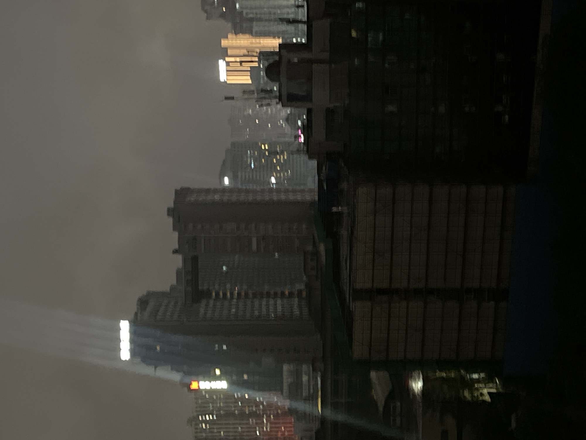 The image depicts a nighttime cityscape in Jiulongpo District, China, under hazy or foggy conditions. Numerous high-rise buildings populate the skyline, with artificial illumination from windows and rooftop signs creating glows against the overcast sky. In the left-center, a prominent skyscraper displays illuminated letters, appearing to spell "SOHO" or similar, on its rooftop. A distinct, upward-angled beam of white light originates from the lower-left foreground and traverses vertically across the upper portion of this building. To the right, other distant high-rises are visible, including one with a striking golden-orange vertical illumination pattern. The immediate foreground consists of dark, indistinct structures, possibly covered buildings or construction sites, exhibiting rectangular patterns consistent with stacked materials or scaffolding. A dark, reflective surface in the lower-left reflects ambient light. No individuals, vehicles, or specific actions are discernible within the frame.