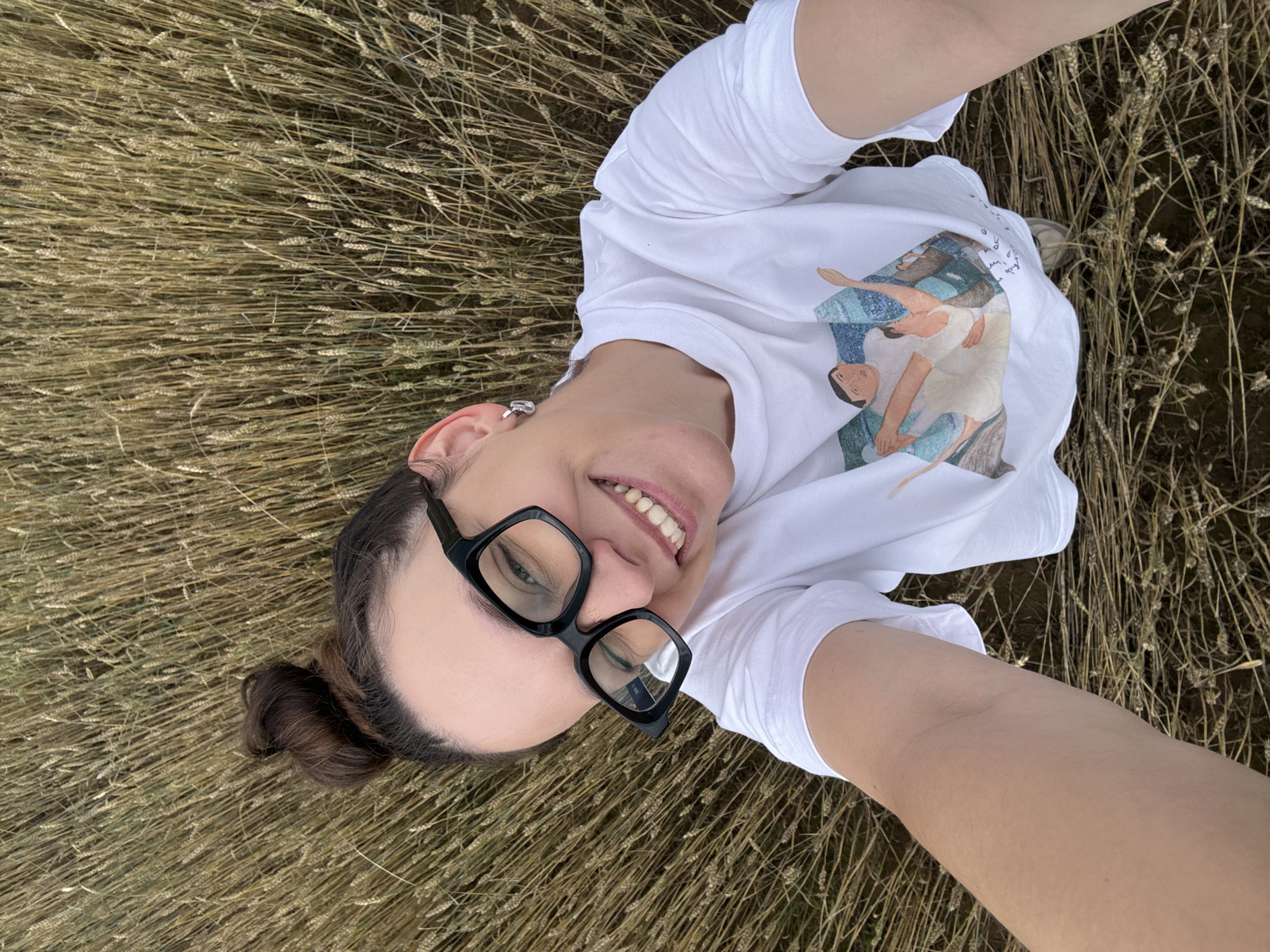 A woman is lying down in a field of wheat, taking a selfie. She is wearing glasses, a white t-shirt with a graphic print, and hoop earrings. Her hair is tied up in a bun. The wheat field is golden brown and appears dry. The location is the city of Lukintsy, Russia.