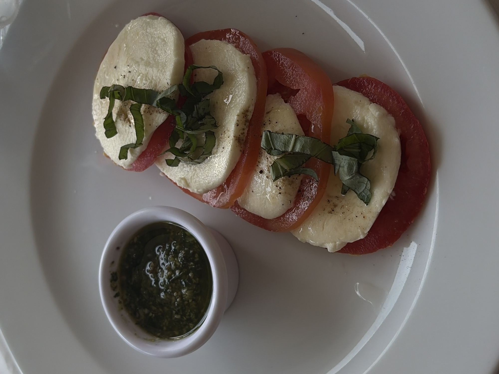 An overhead, close-up shot displays a Caprese salad served on a clean, round white ceramic plate. The main subjects are alternating slices of fresh mozzarella cheese and red tomatoes, arranged in an overlapping diagonal line across the plate. Each stack is garnished with fresh green basil leaves. A light drizzle of olive oil and a sprinkle of what appears to be black pepper or dried herbs are visible on the cheese and tomatoes.
To the lower left of the salad, a small, white ceramic ramekin holds a vibrant green sauce, likely pesto, which shows some texture and gloss. The scene depicts a prepared meal, possibly an appetizer, ready for consumption in a dining setting. The lighting is soft and even, without harsh shadows, suggesting an indoor environment or diffused natural light. No people are visible, and there are no discernible emotions conveyed. No text is present in the image. While the context of Puebla, Mexico, is provided, there are no specific visual cues within the photograph to indicate this location.