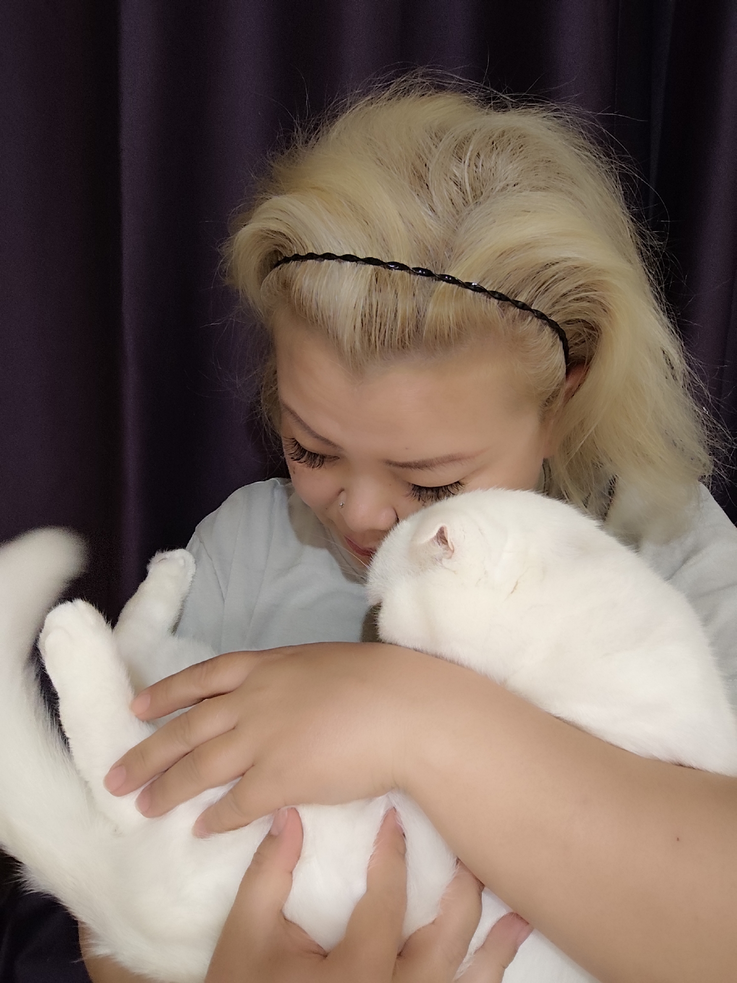 A person with blonde hair and a black headband is holding a white cat. The person is wearing a light-colored shirt. The background appears to be a dark curtain. The location is unknown.